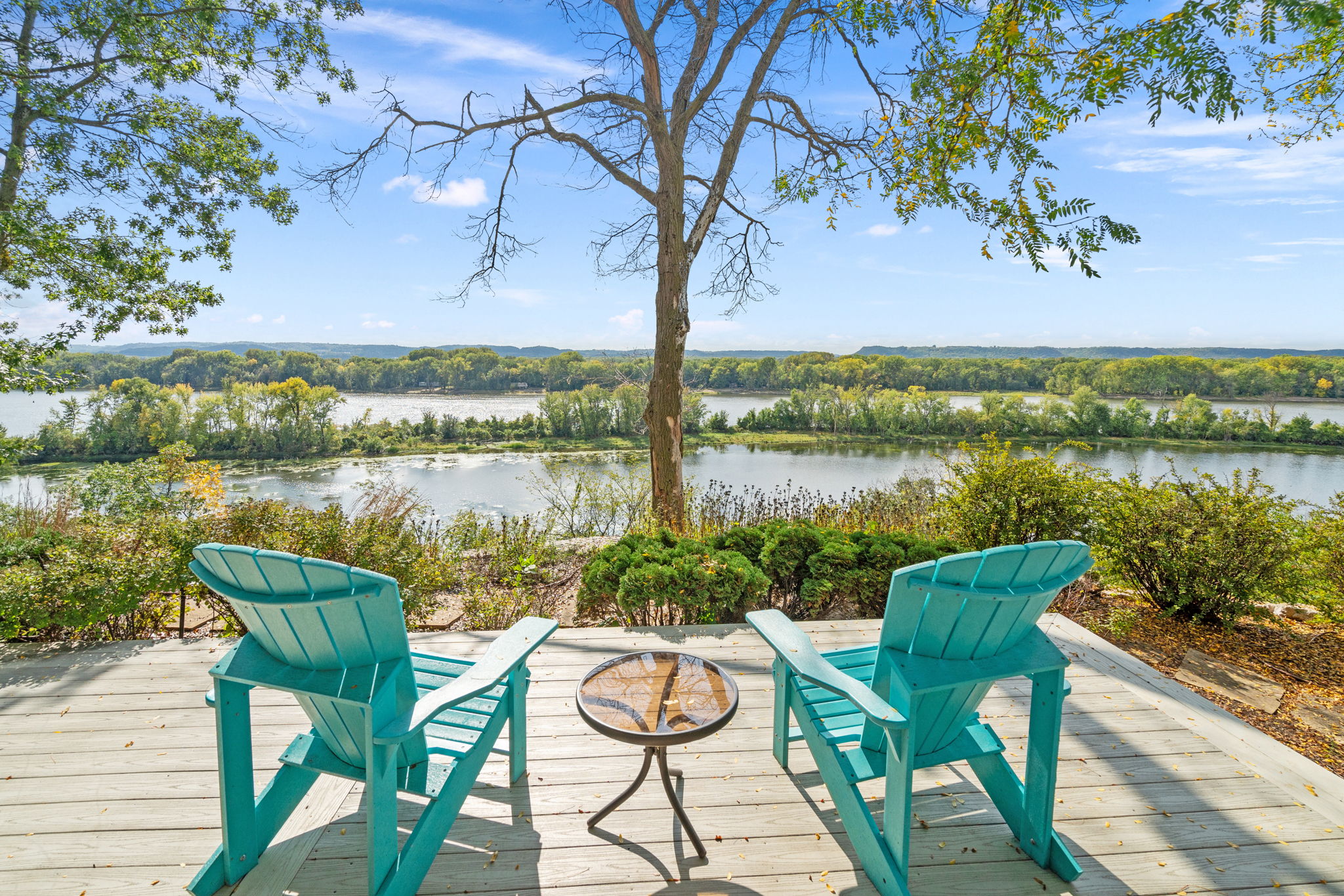 A tranquil riverside view featuring turquoise Adirondack chairs and autumn foliage.