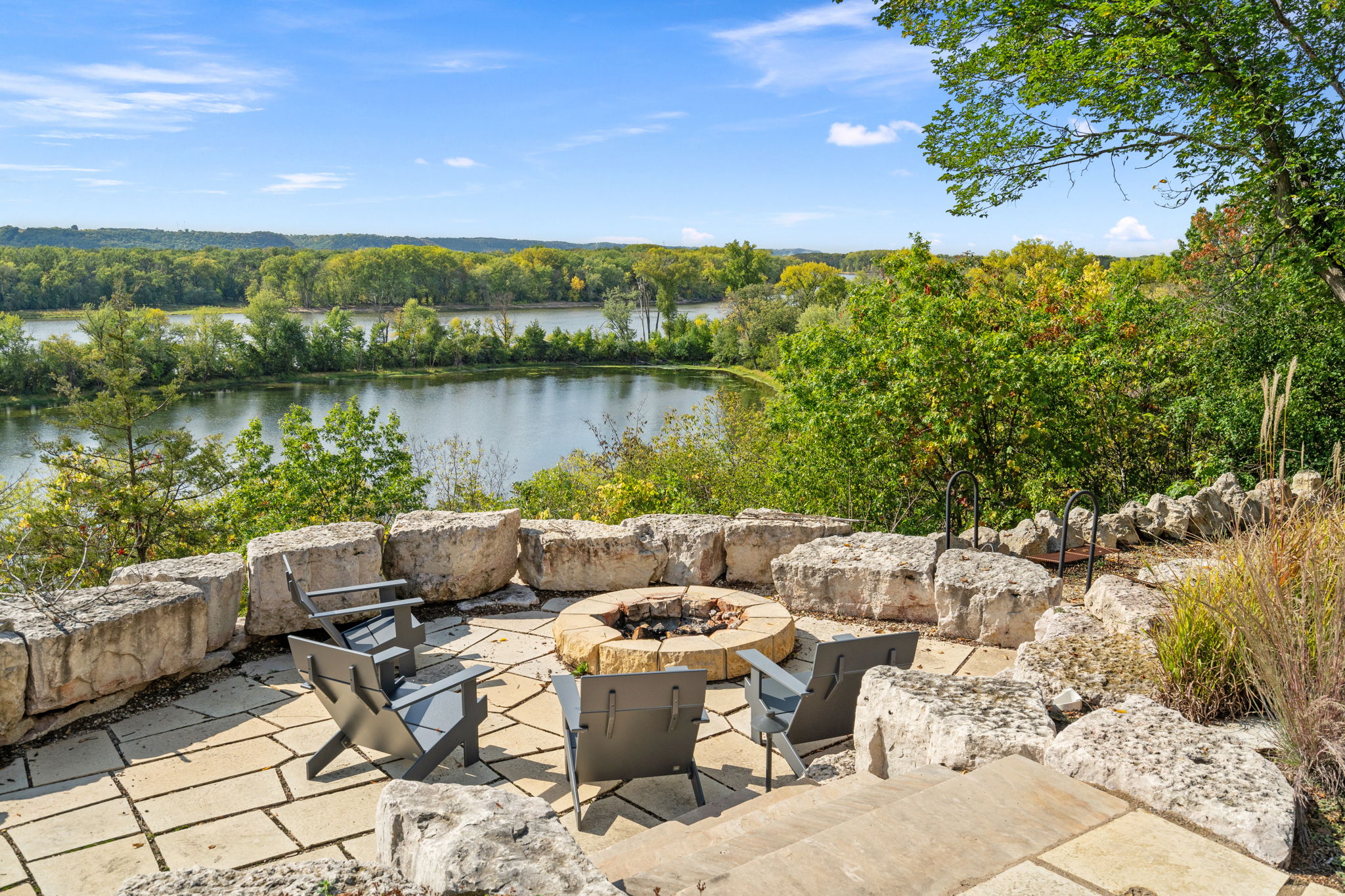 A cozy outdoor fire pit area with chairs, overlooking a scenic river and lush greenery.