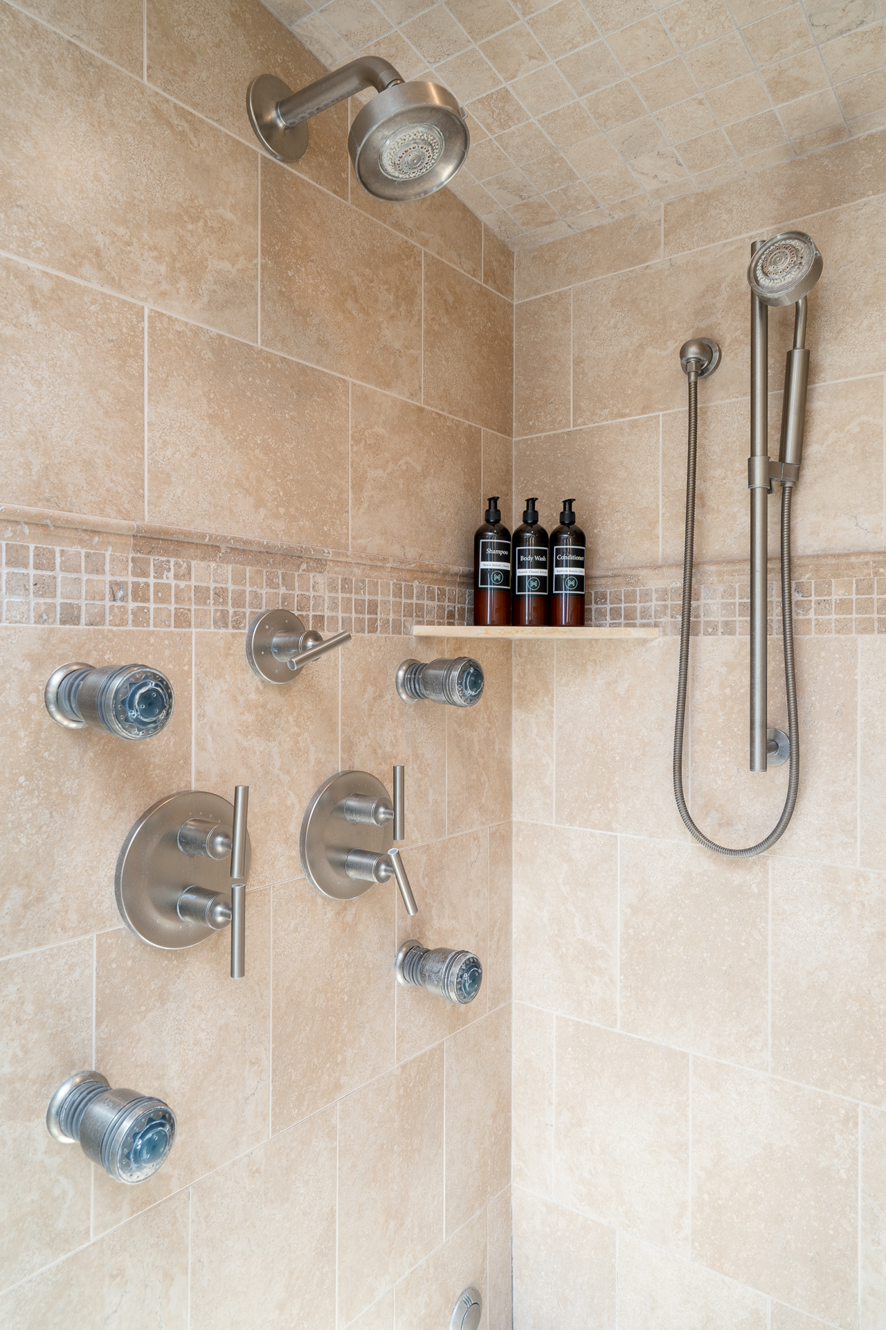 A modern shower with beige tiles and sleek chrome fixtures, featuring stylish dark glass bottles on a wooden shelf.