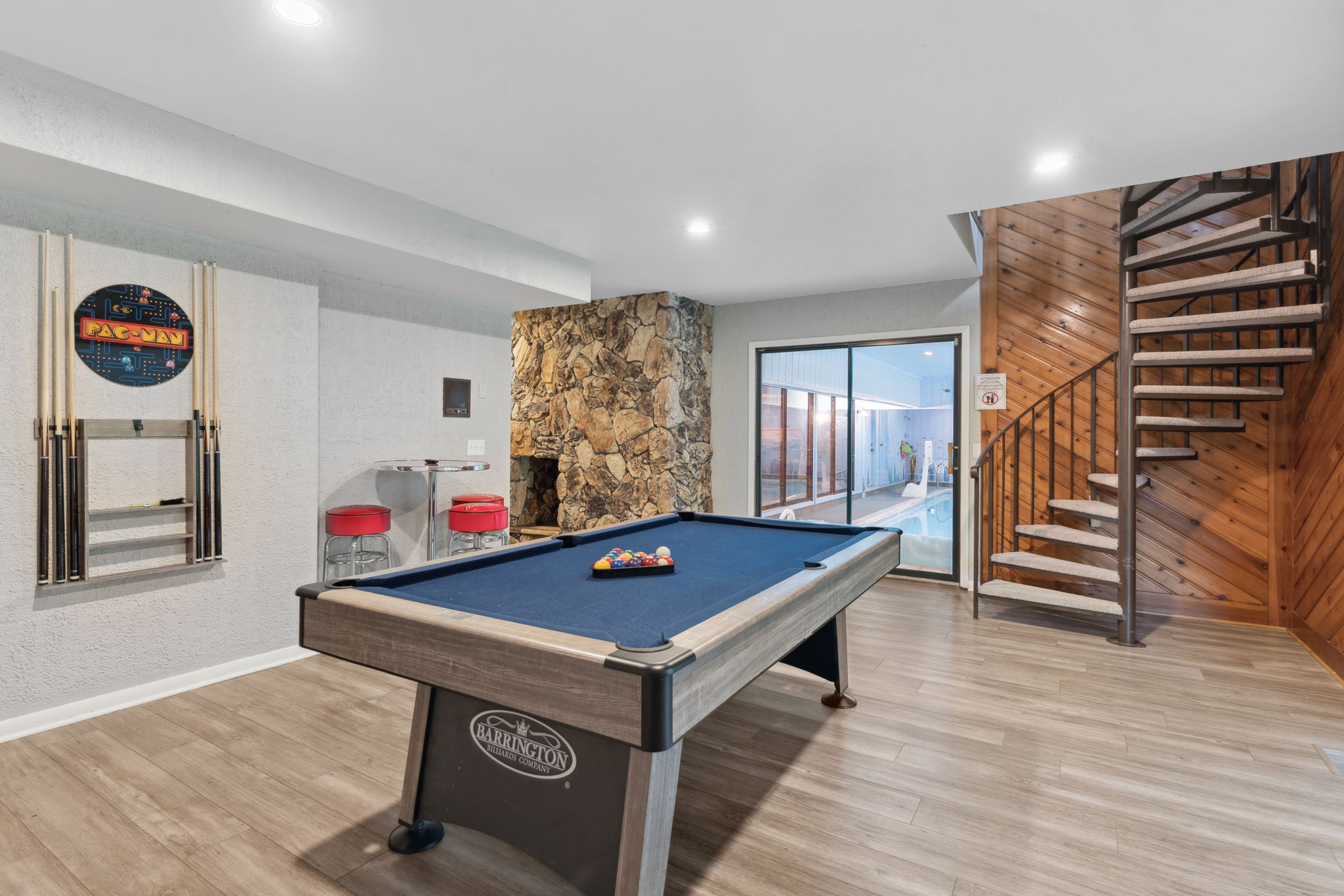 A stylish game room featuring a billiard table, arcade sign, and a view of an indoor pool.