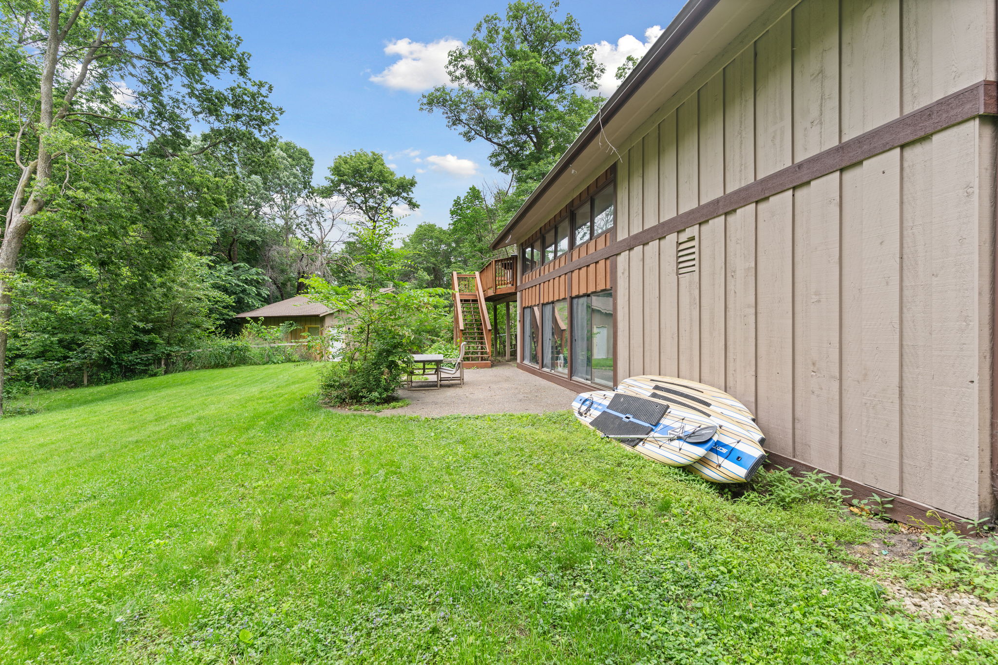 A serene backyard view featuring a two-story house and paddleboards, surrounded by lush greenery.