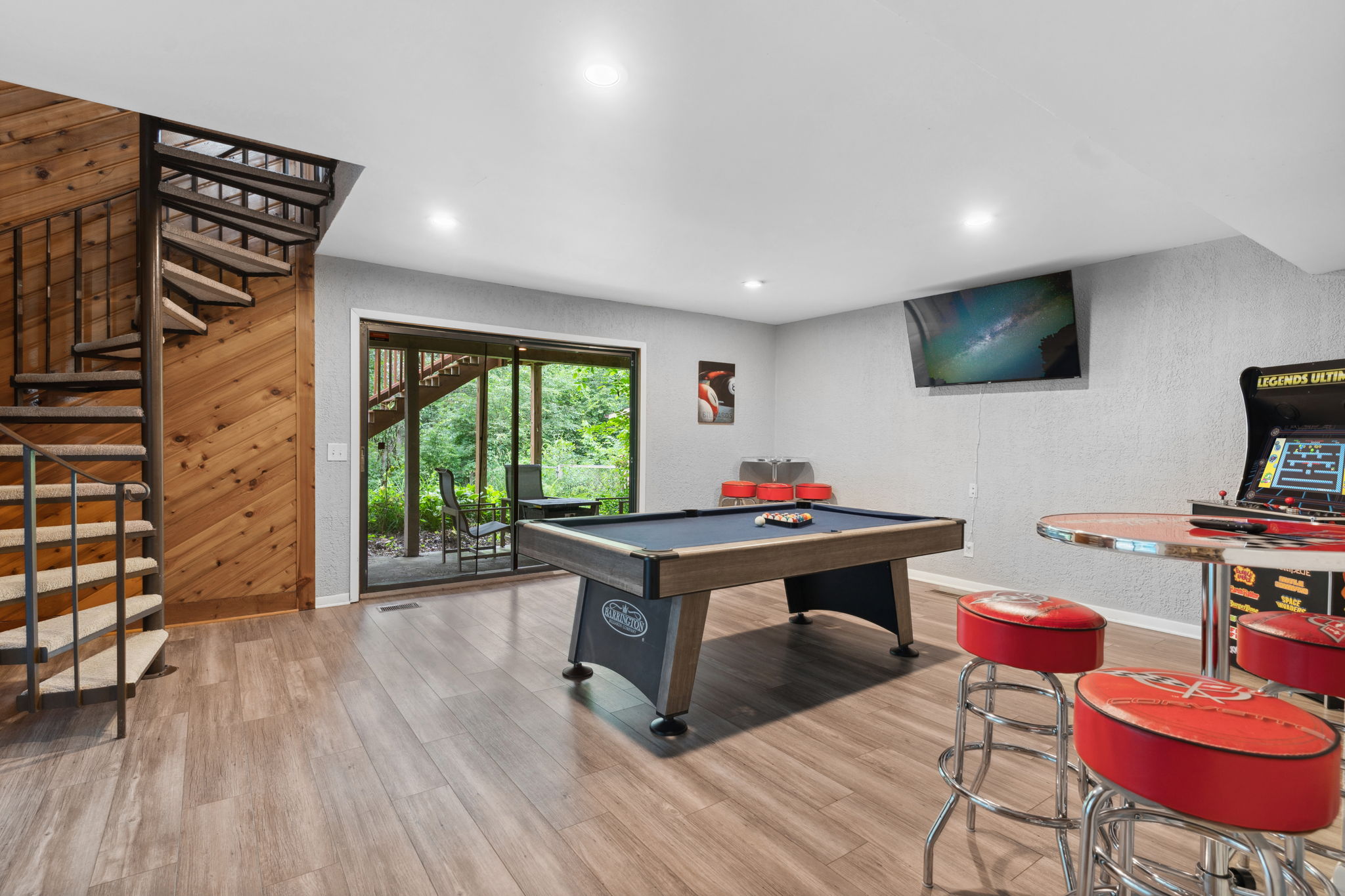 A cozy entertainment room featuring a pool table, arcade game, and sliding door to a green patio.