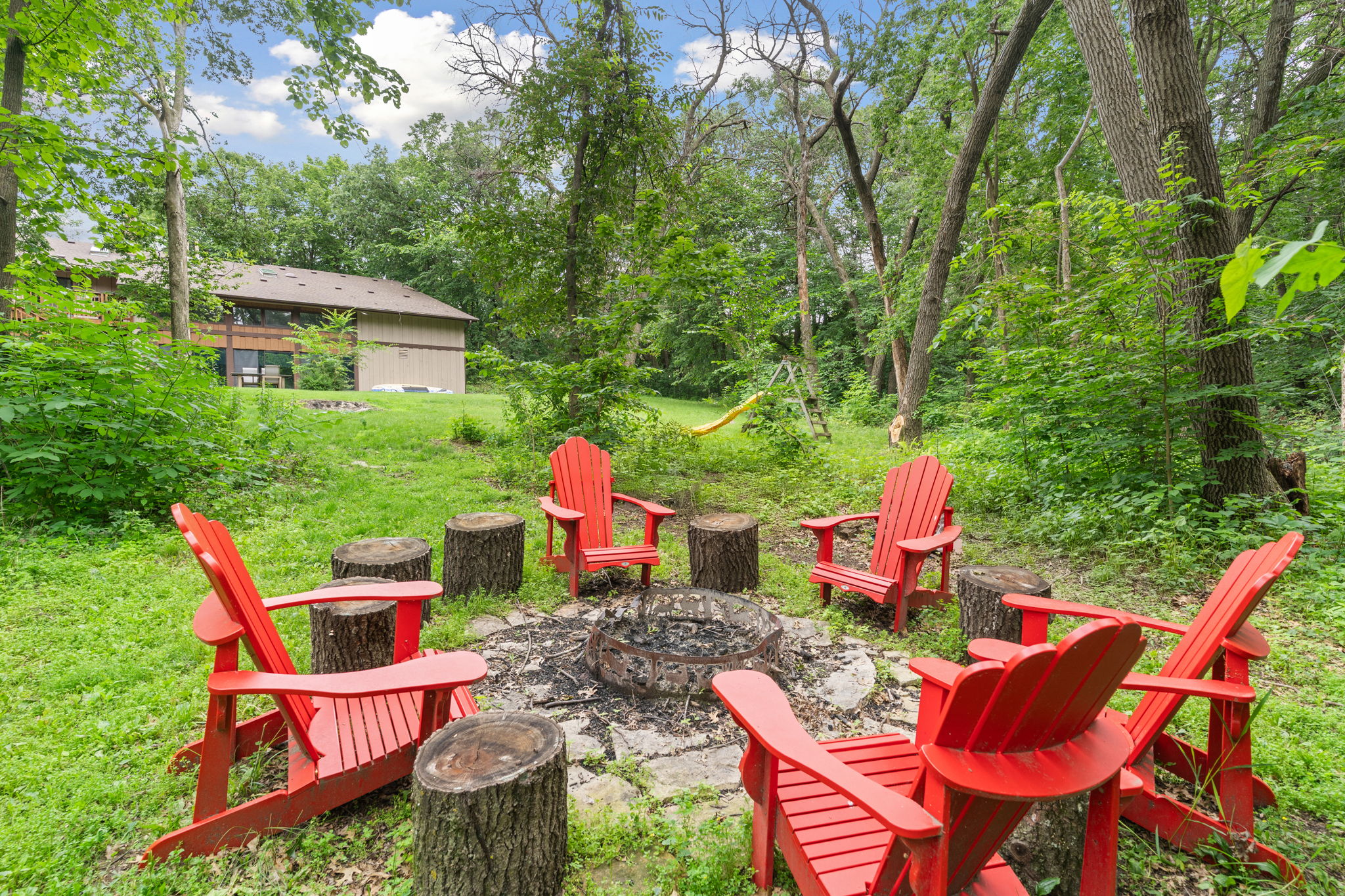 A cozy outdoor fire pit surrounded by red Adirondack chairs in a lush green setting.