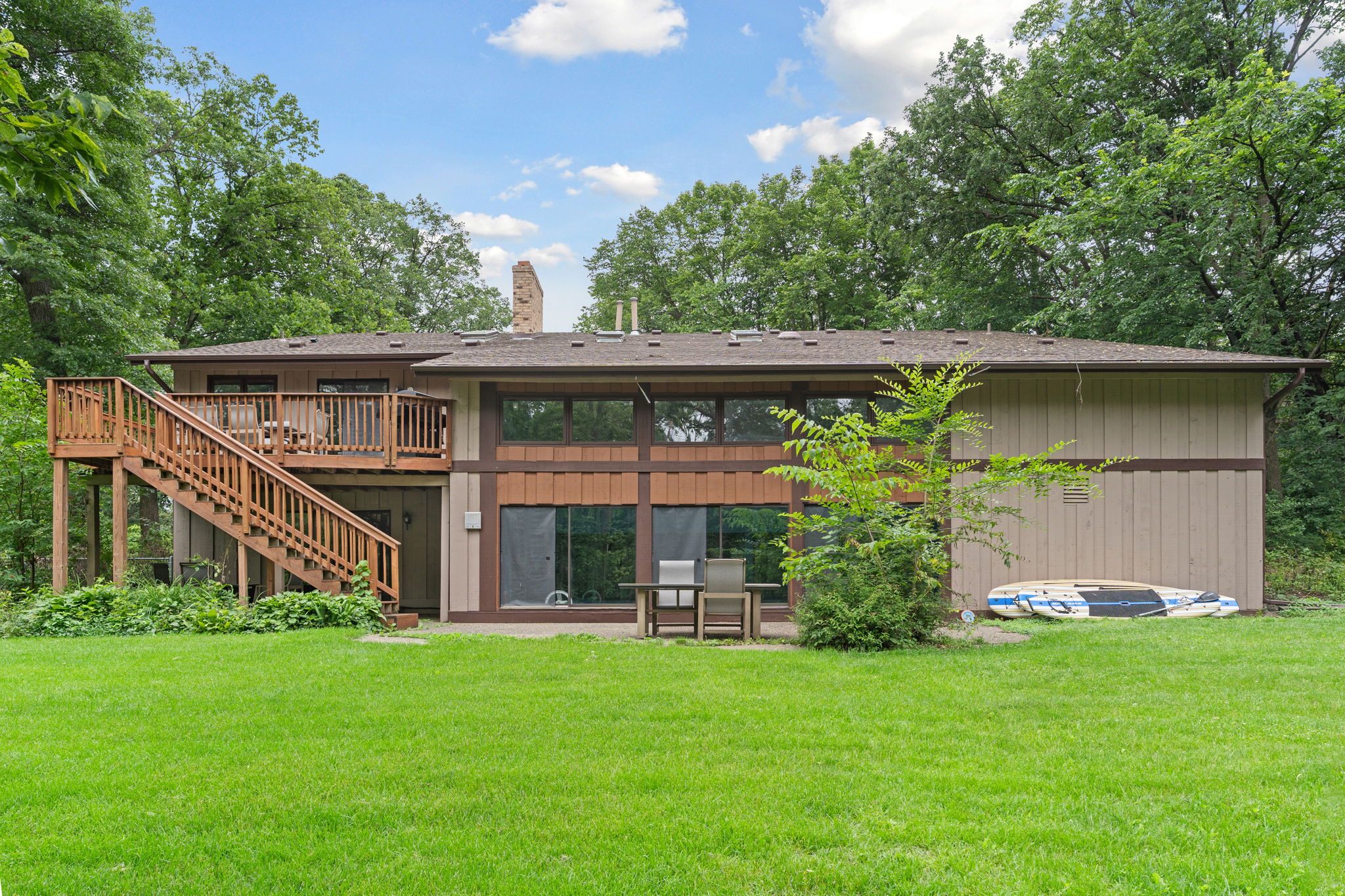 Charming two-story house surrounded by lush greenery, featuring a wooden deck and patio seating.