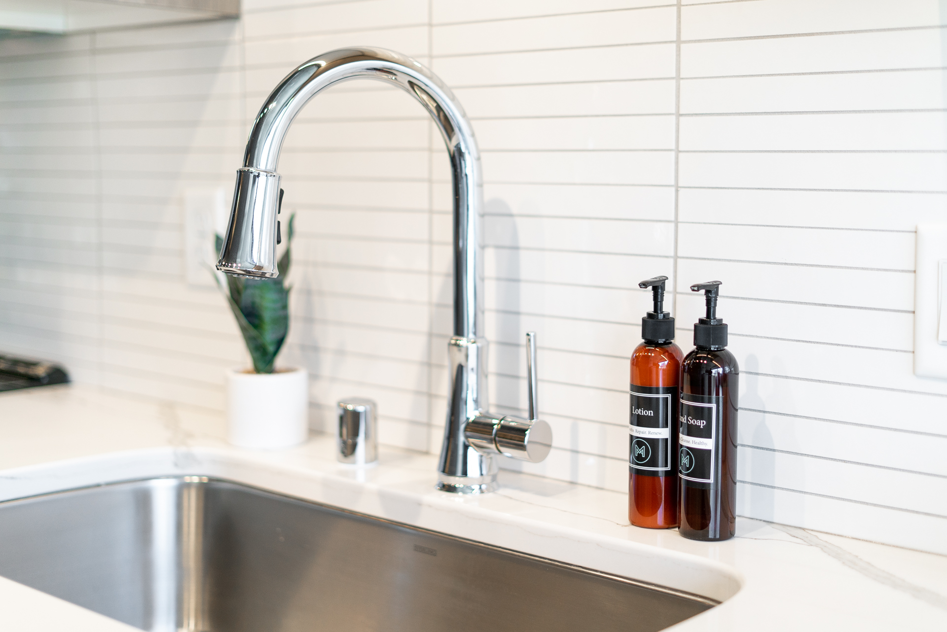 A modern kitchen sink area featuring a sleek faucet and labeled lotion and hand soap bottles.