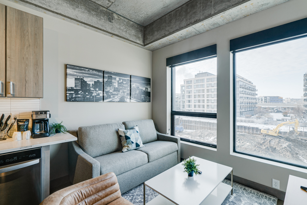 Modern living room with gray sofa and kitchen area, featuring city skyline art and construction view outside.