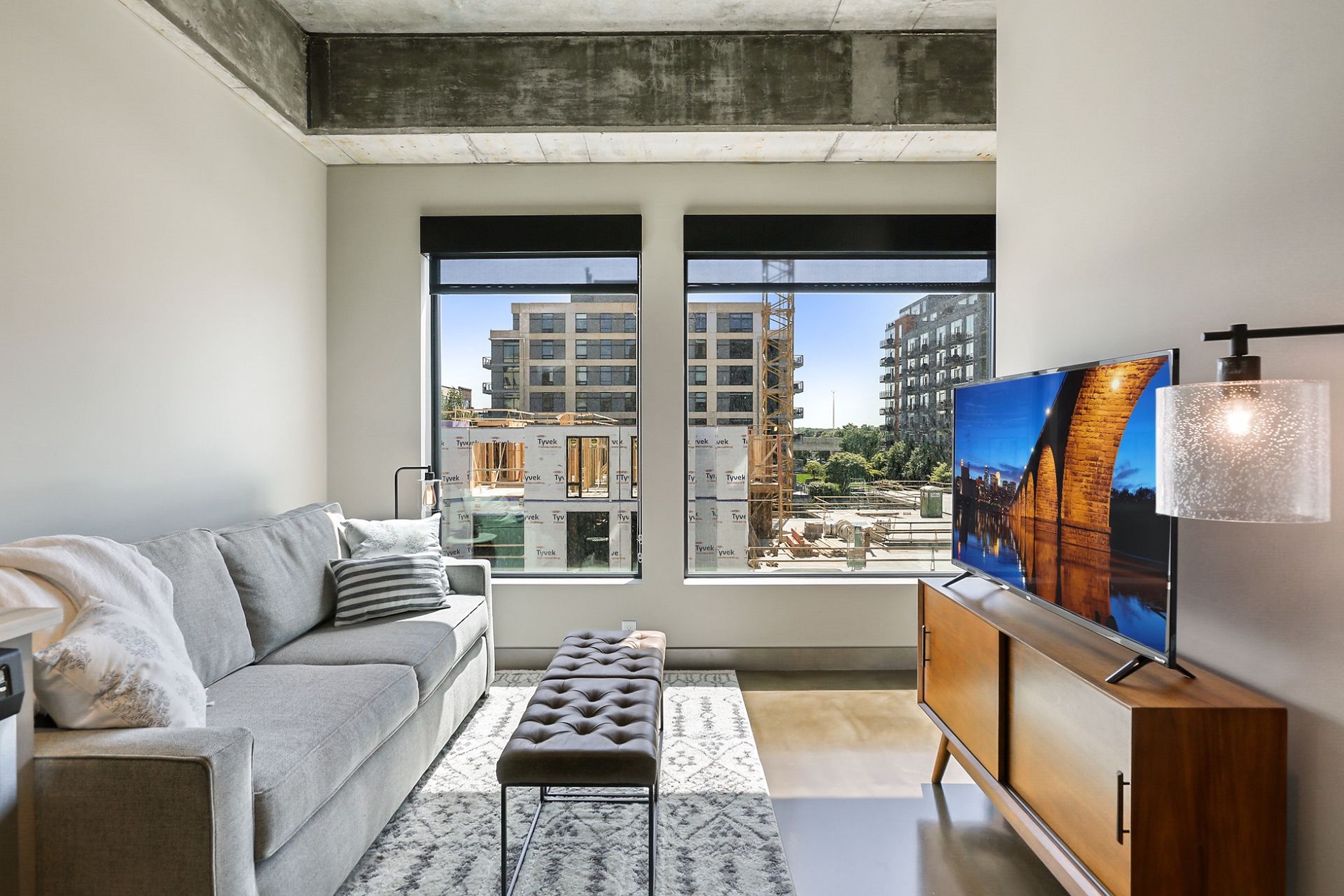 Modern living room featuring a grey sofa, a tufted ottoman, and large windows overlooking a construction site.