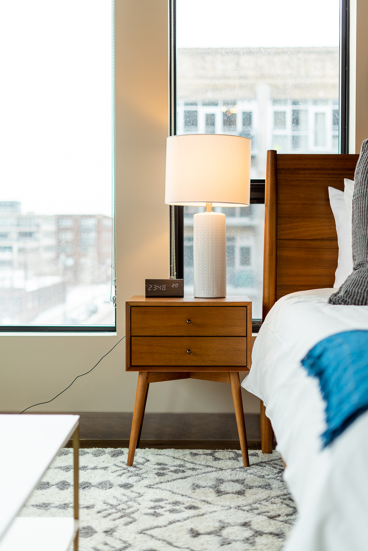 A modern nightstand with a lamp and digital clock beside a cozy bed in a rainy city bedroom.