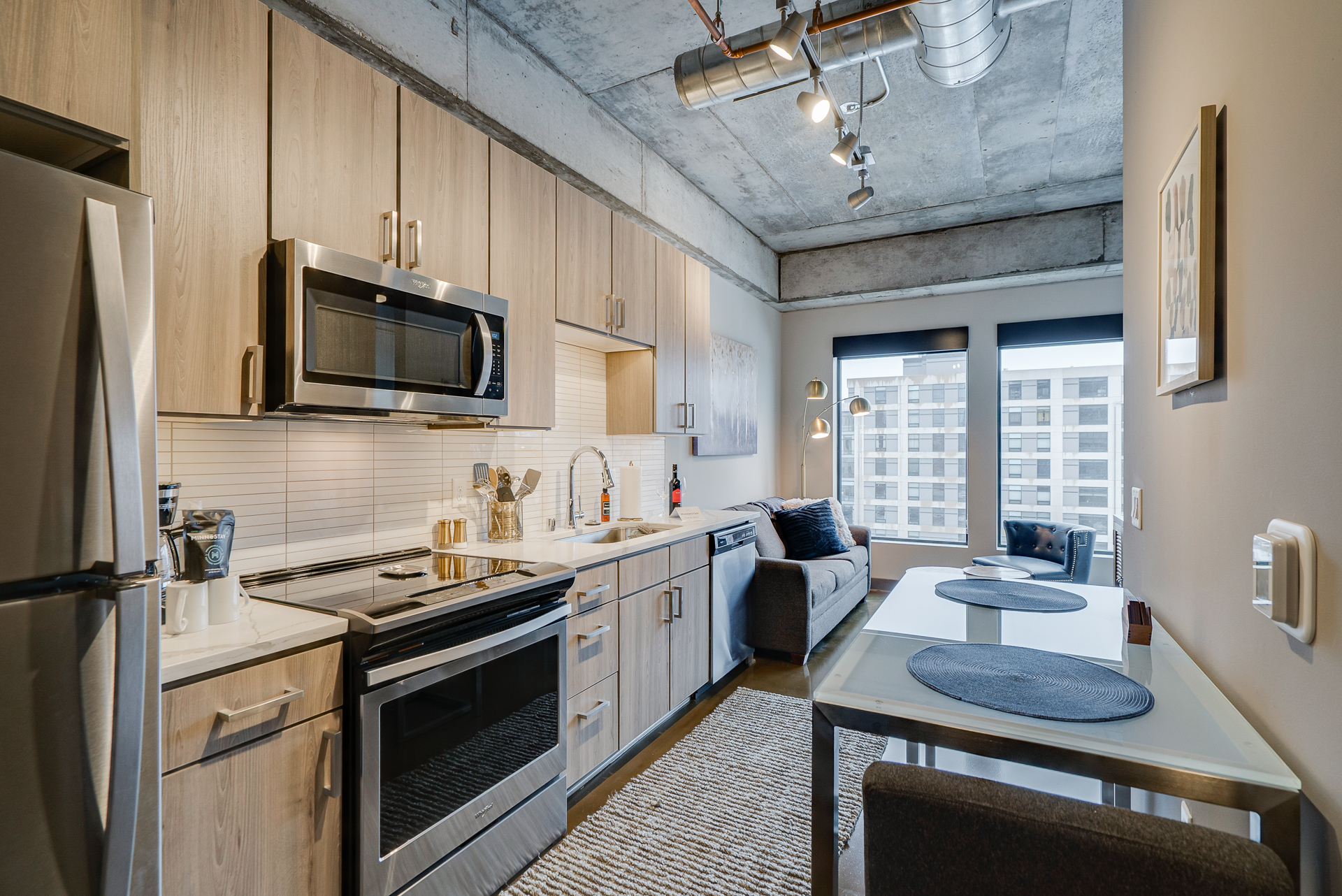 Modern kitchen with light wood cabinets, stainless steel appliances, and a cozy living area with large windows.