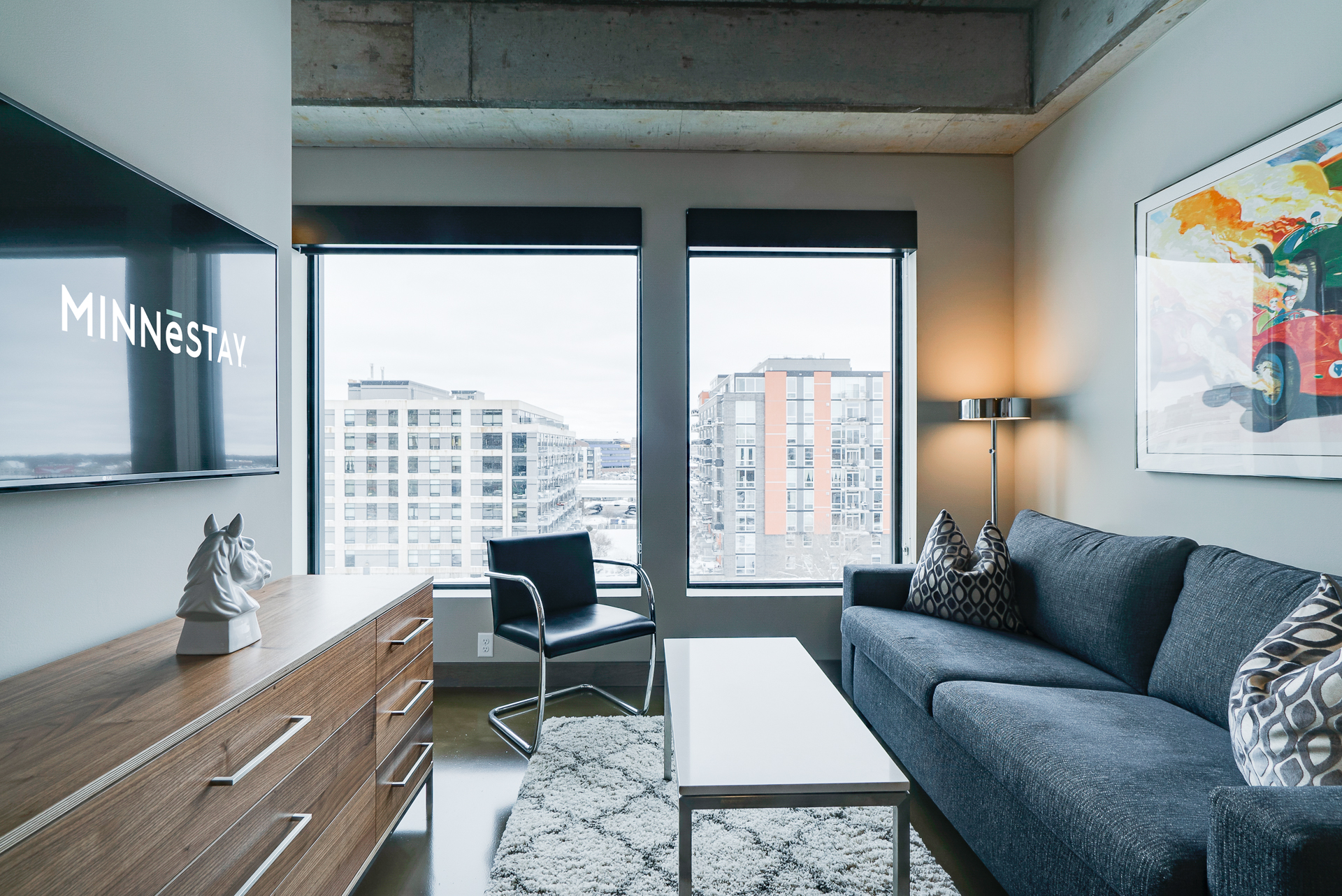 Modern living room with a sofa, artwork, and large windows showing the city.