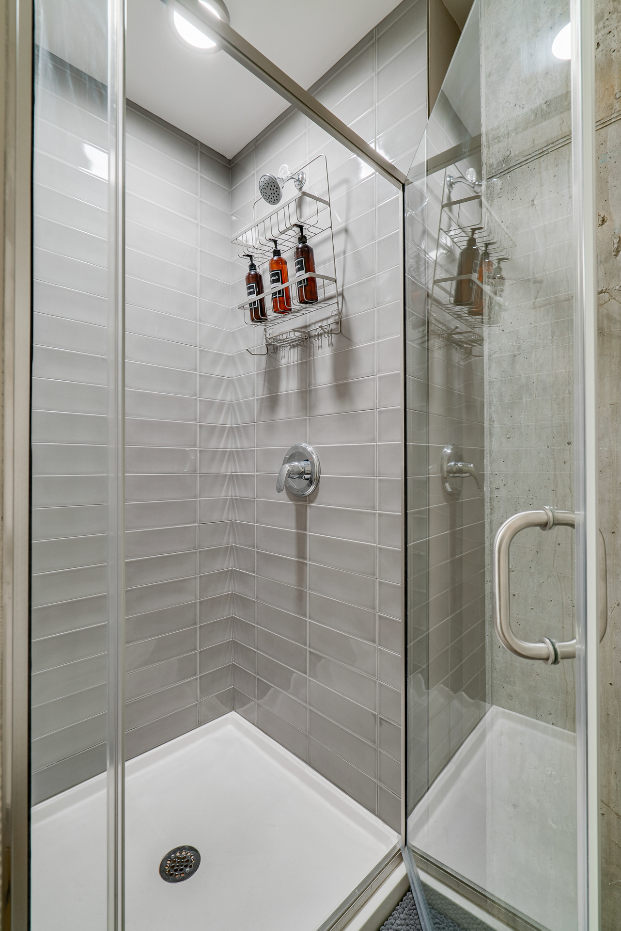 Modern shower enclosure with gray tiles and a silver shelving unit for products.