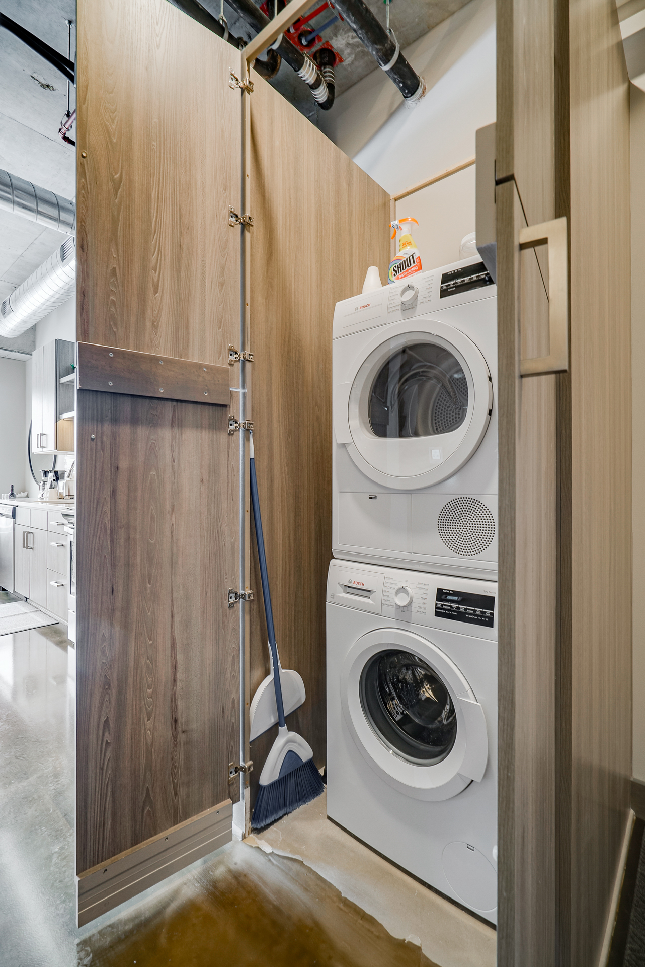 A modern laundry nook featuring a stacked washer and dryer, tucked behind a wooden panel with cleaning supplies nearby.