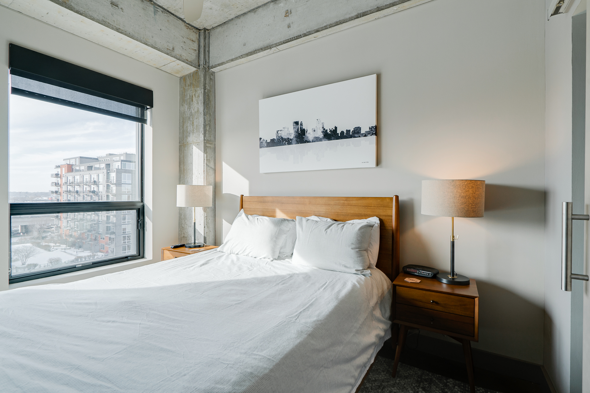 A modern bedroom with a minimalist design, showcasing a wooden bed, abstract artwork, and bright natural light.
