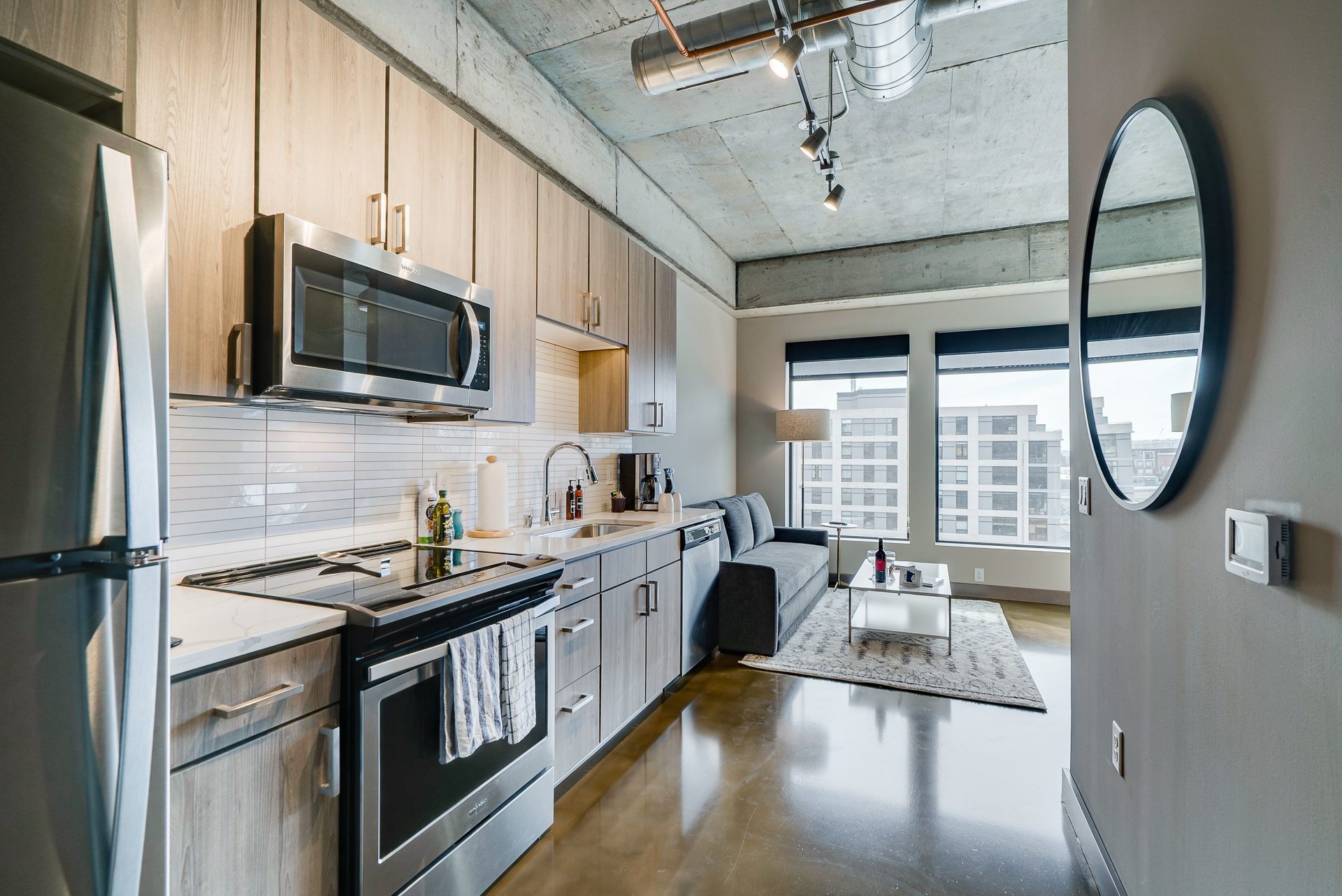 Interior view of a contemporary kitchen and living area in an apartment, featuring stainless steel appliances and a gray sofa.