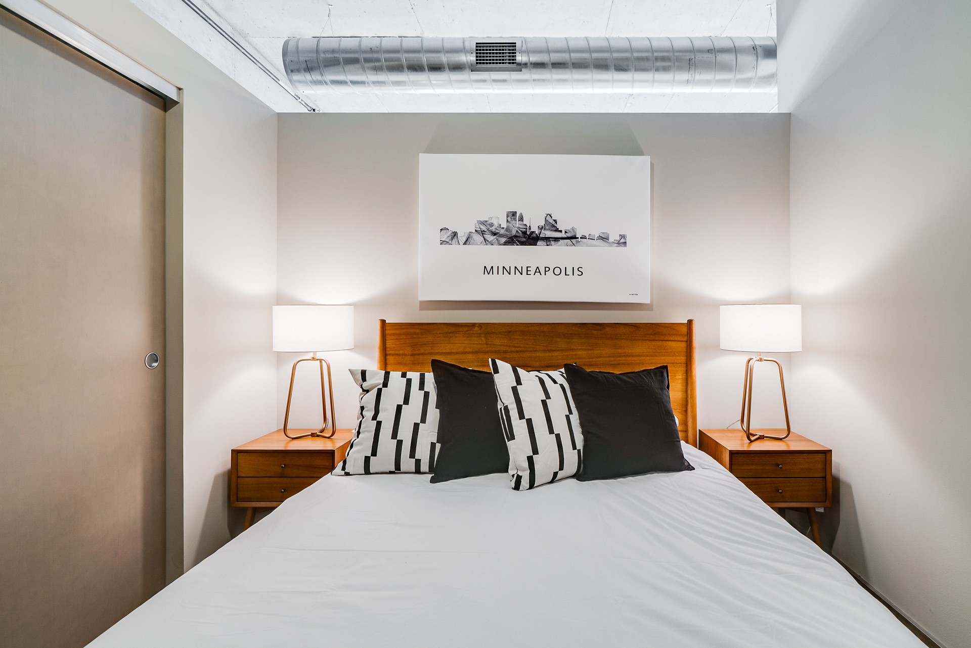 A modern bedroom featuring a minimalist design with a bed, stylish pillows, and a skyline artwork.