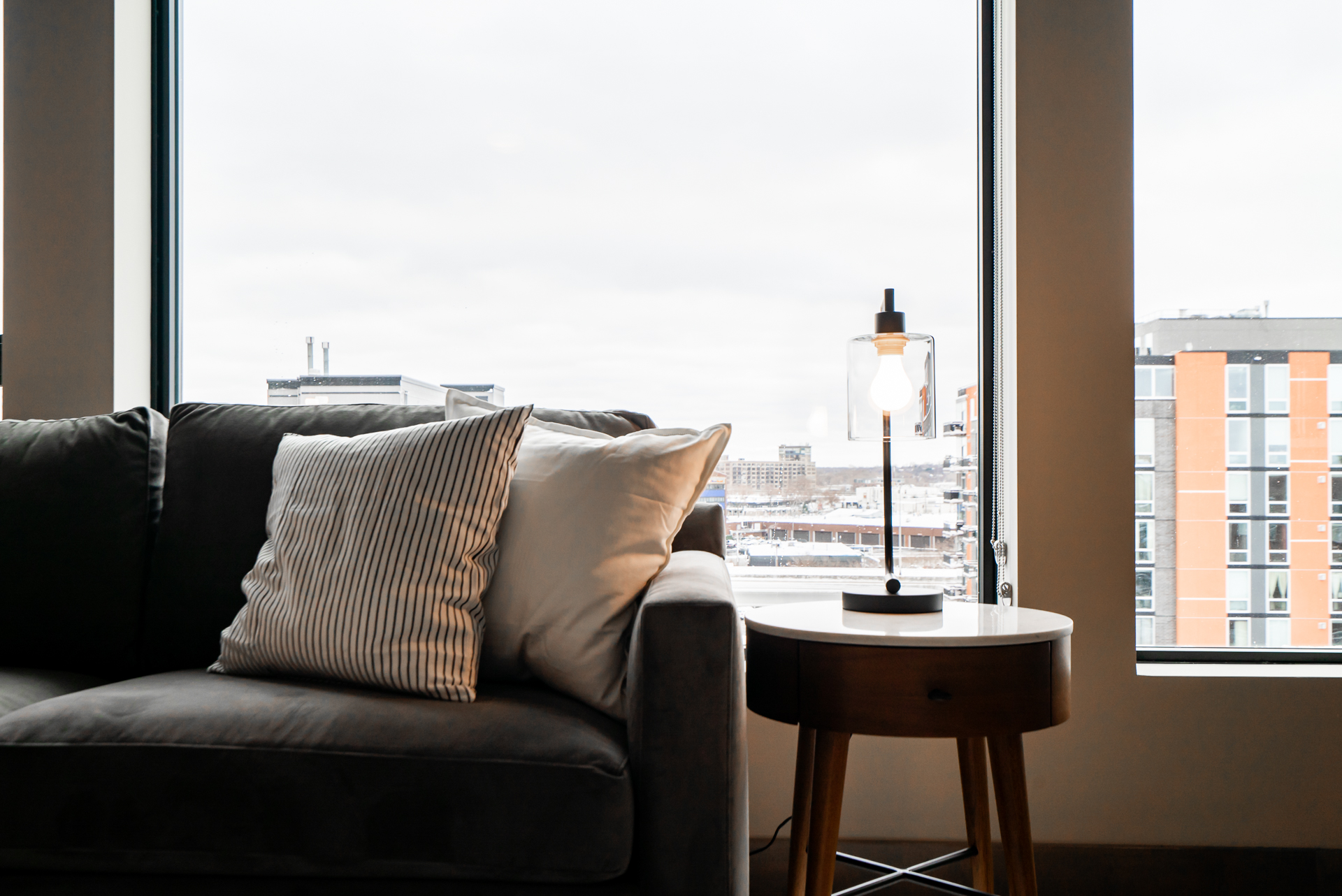 A cozy living room corner featuring a stylish gray sofa, modern lamp, and a view of the urban landscape outside.