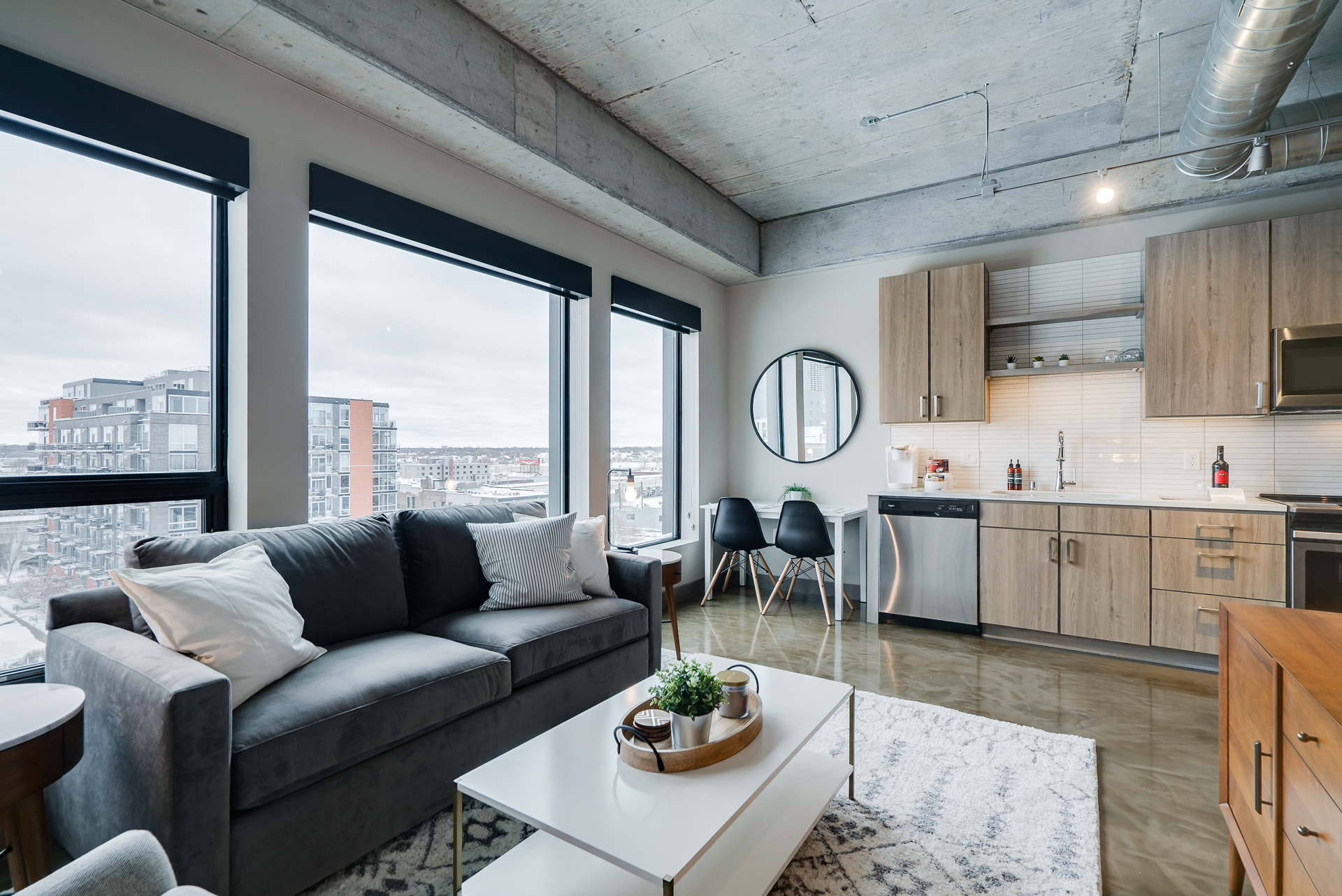 Interior view of a modern apartment with large windows, gray sofa, and a well-equipped kitchen.