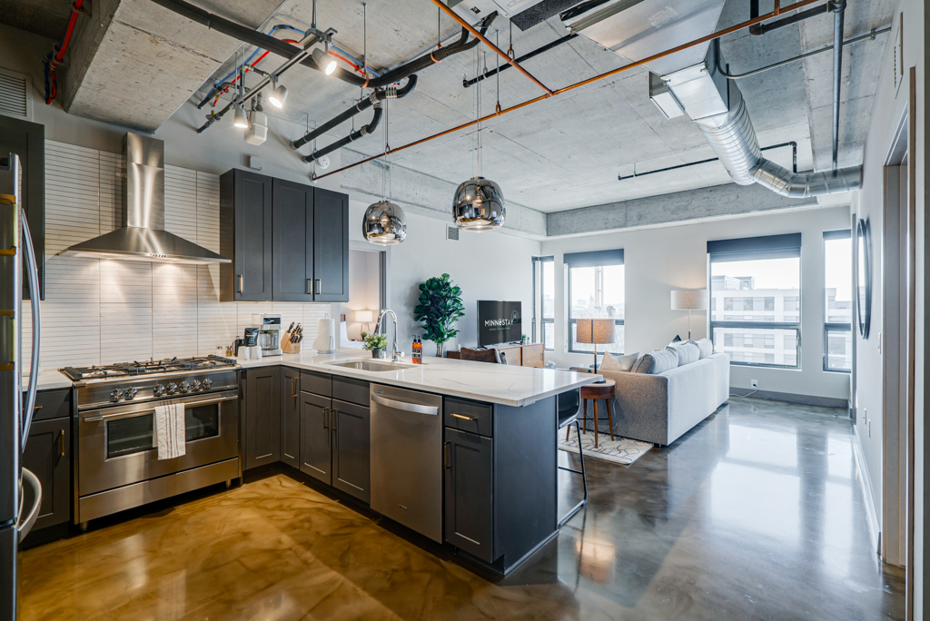 An industrial-style kitchen and living room featuring a sleek design, dark cabinets, and large windows.