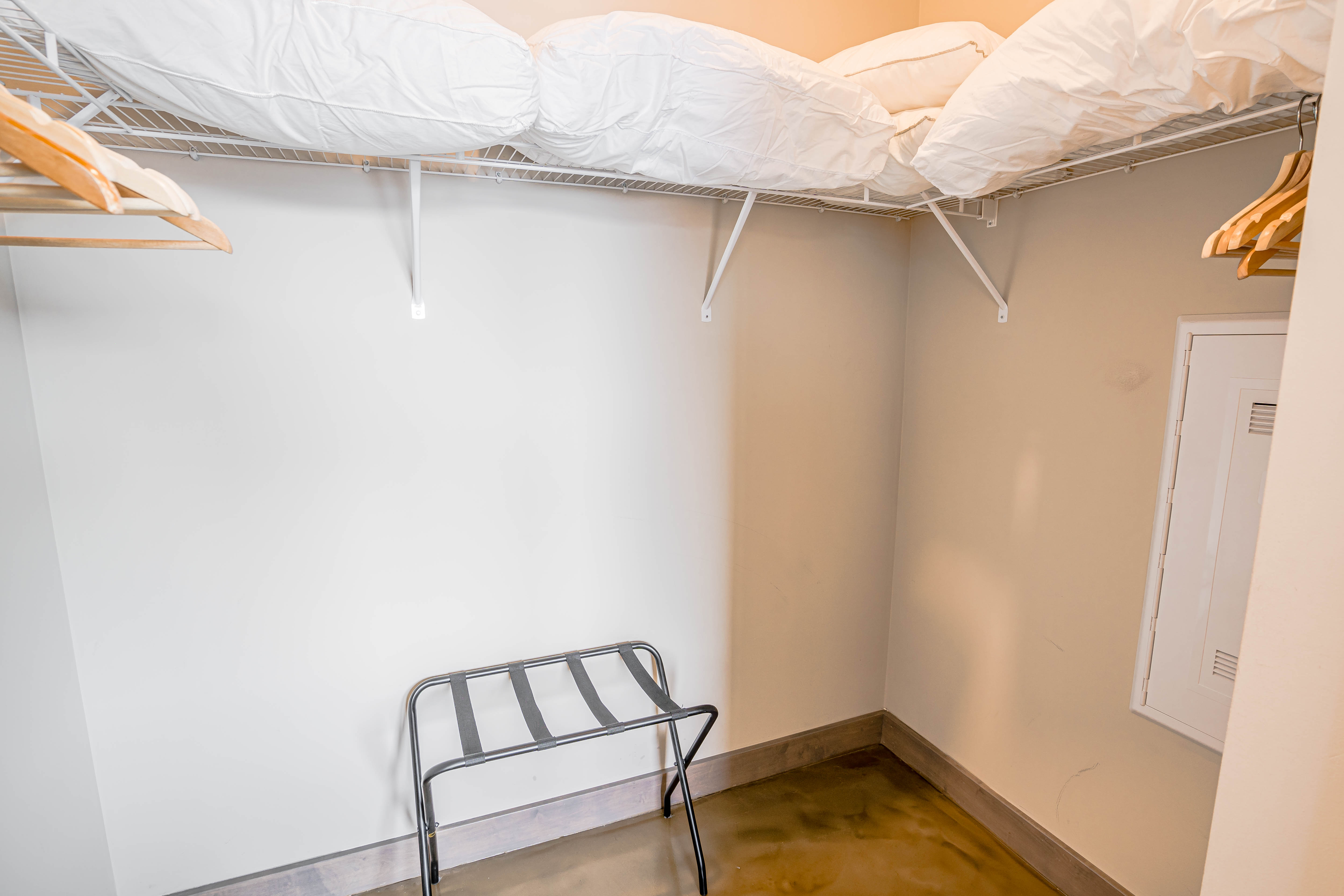A neat closet featuring a wire shelf, white bedding, wooden hangers, and a folding stool.