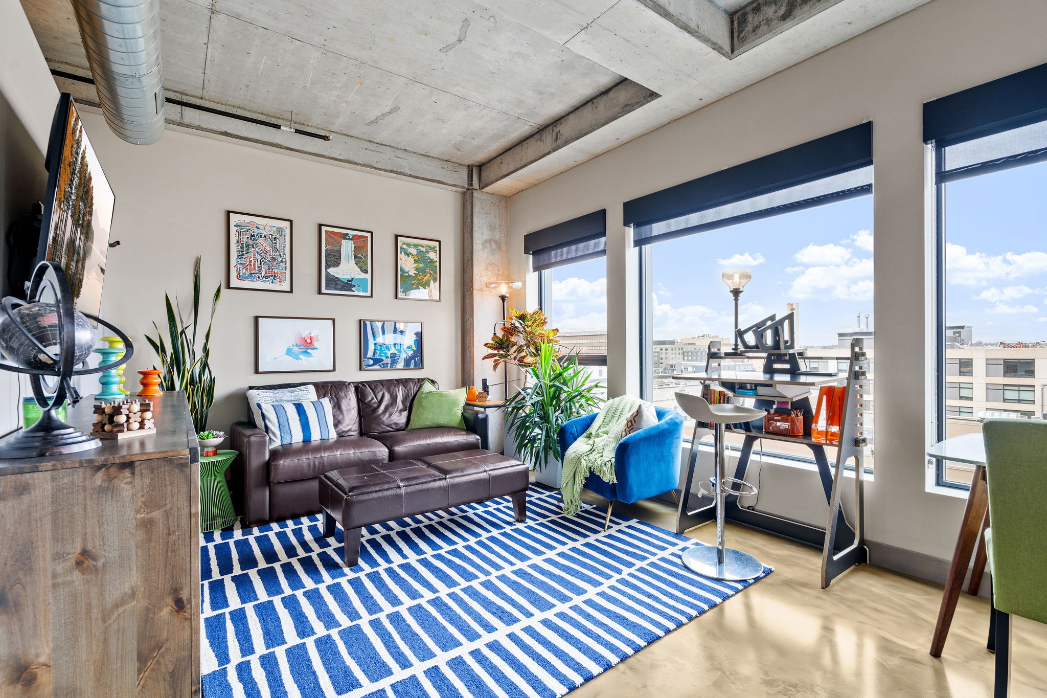 A stylish, modern living room featuring a cozy couch, vibrant artwork, and a workspace with an urban view.