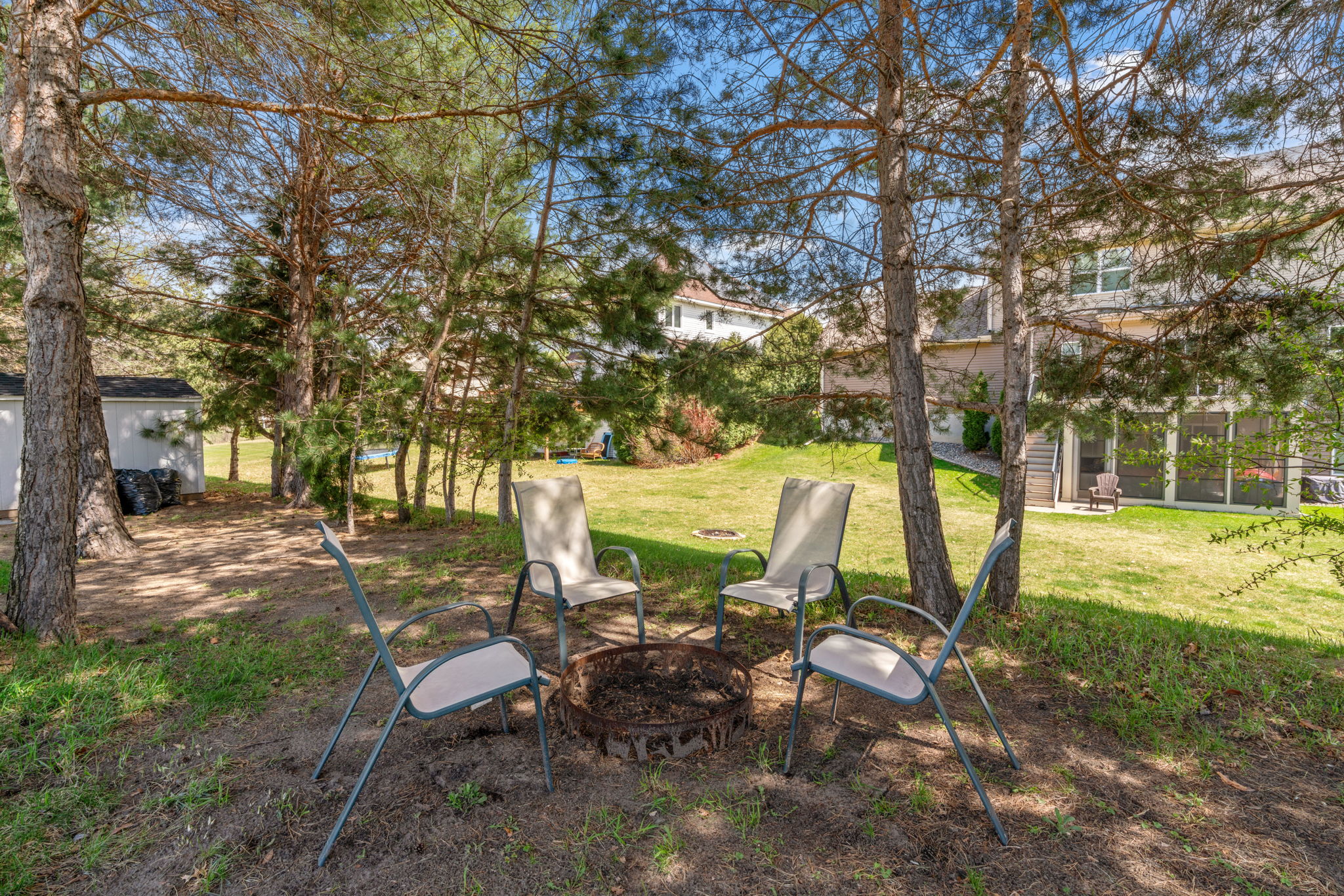 A cozy fire pit area in a sunny backyard, surrounded by trees and lawn.