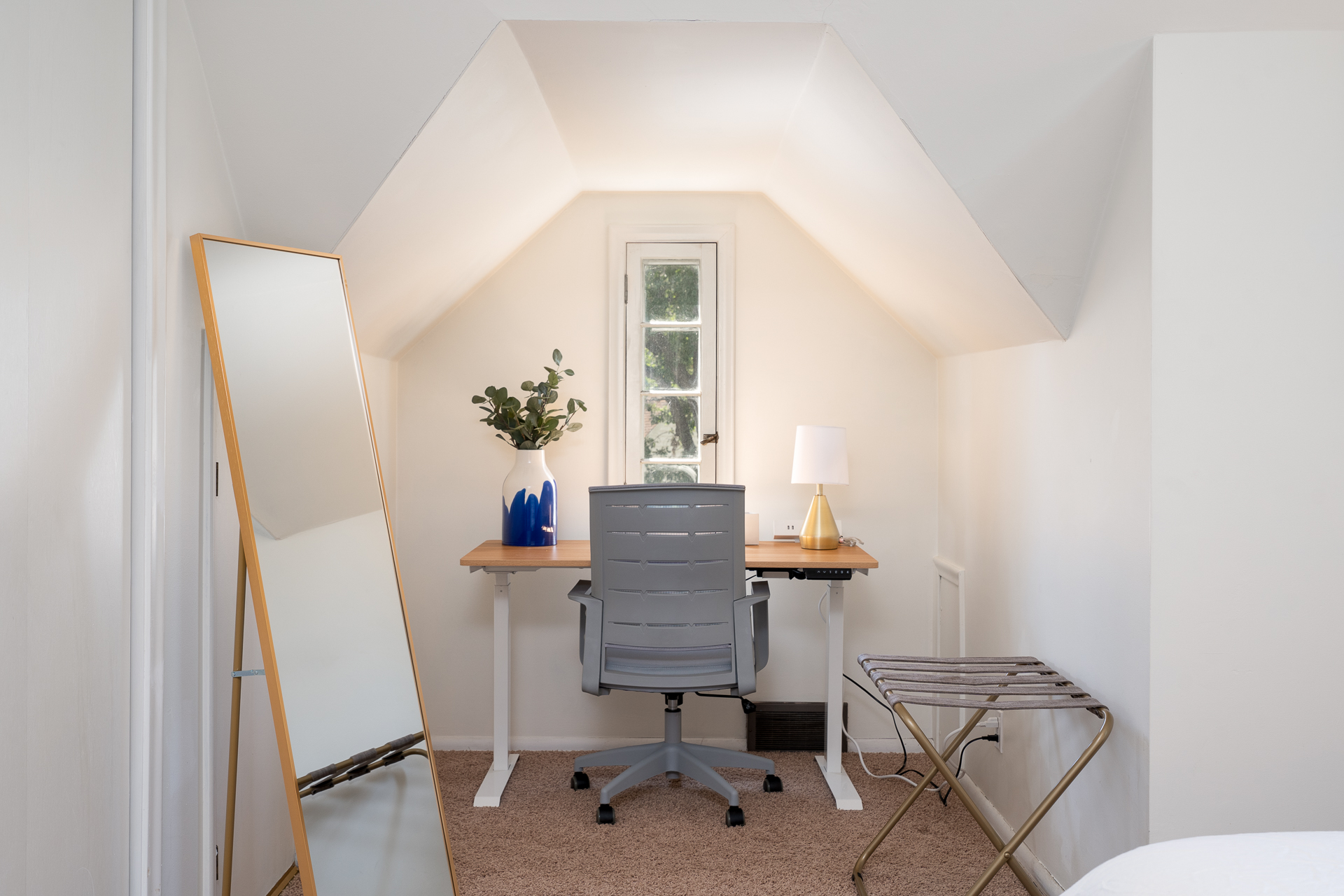 A cozy workspace under a sloped ceiling featuring a wooden desk, a modern chair, and a tall mirror.