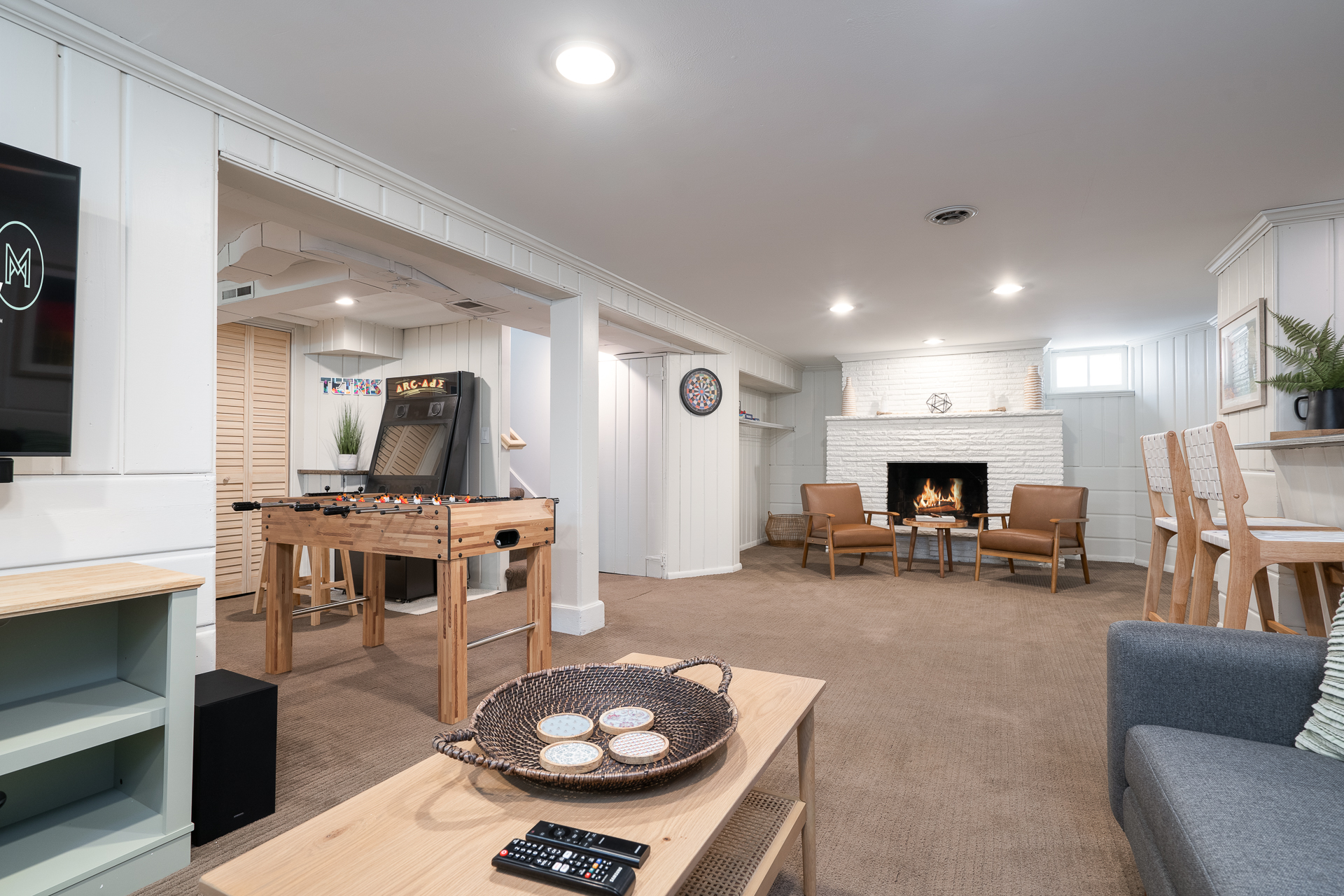 A stylish modern basement living space with a foosball table, arcade game, and a cozy fireplace.