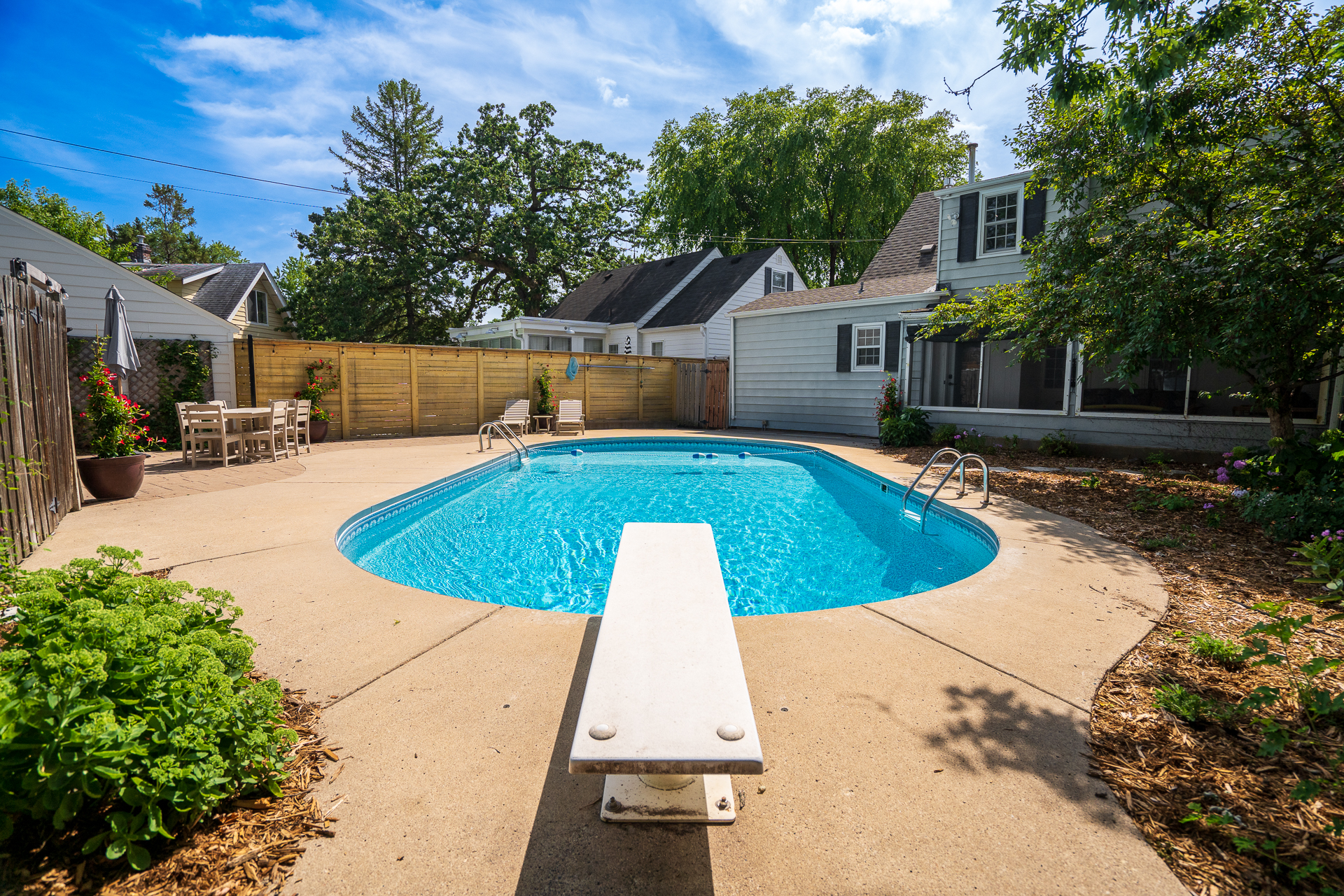 A tranquil backyard pool area with a diving board, surrounded by greenery and cozy seating.