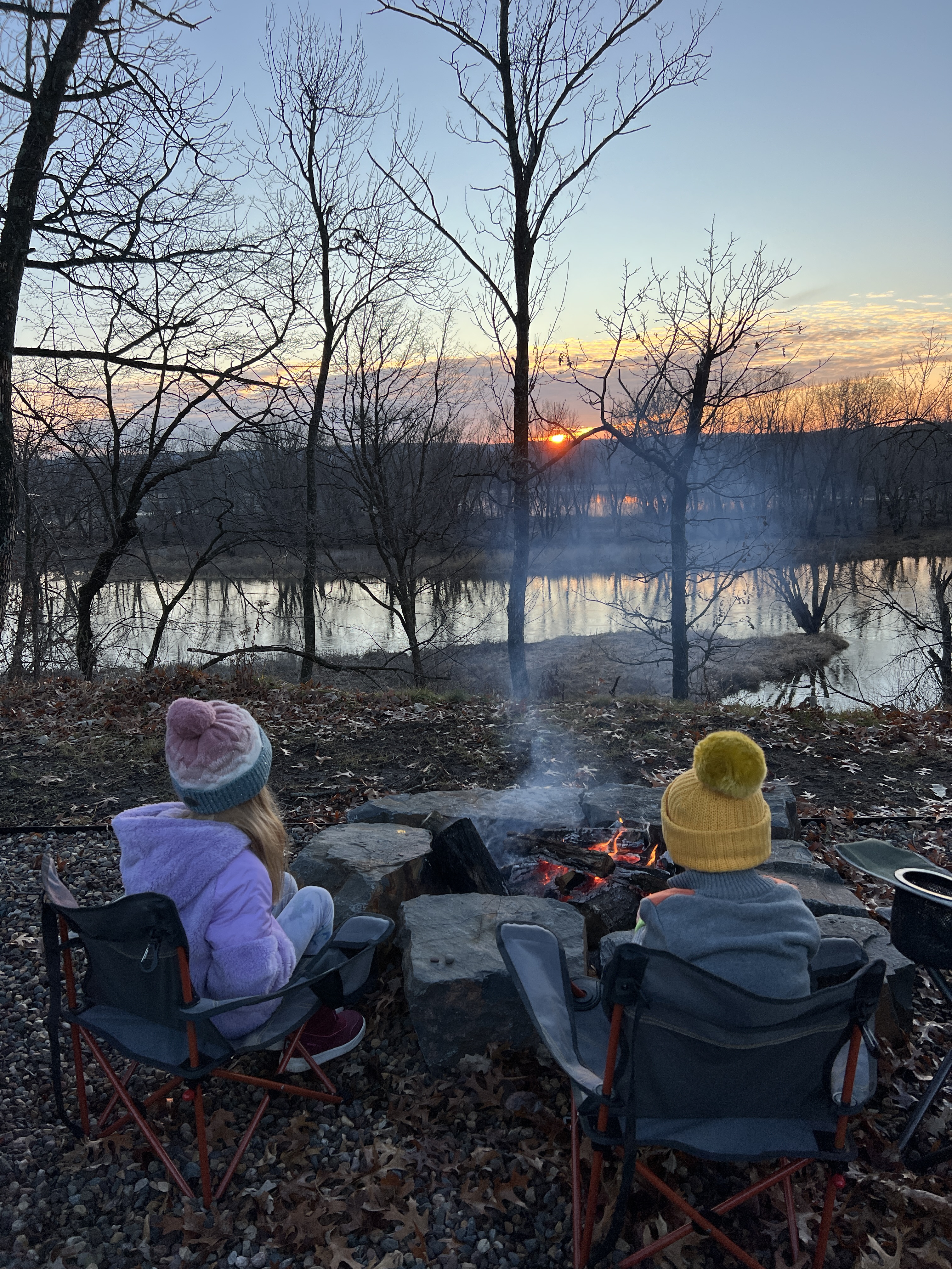 Two children enjoying a campfire at sunset by the lake.