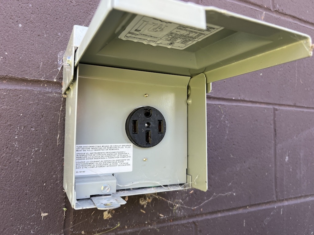 Open electrical outlet housing mounted on a brick wall with a safety instruction label.