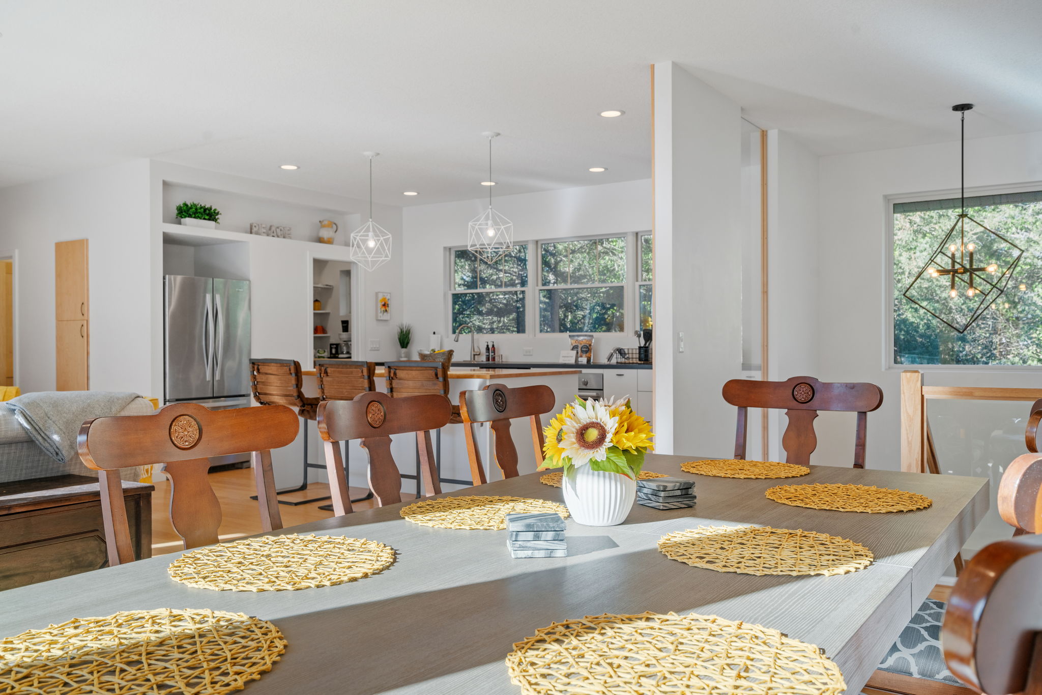 A modern dining area featuring a wooden table with woven placemats and sunflowers, leading into a stylish kitchen.