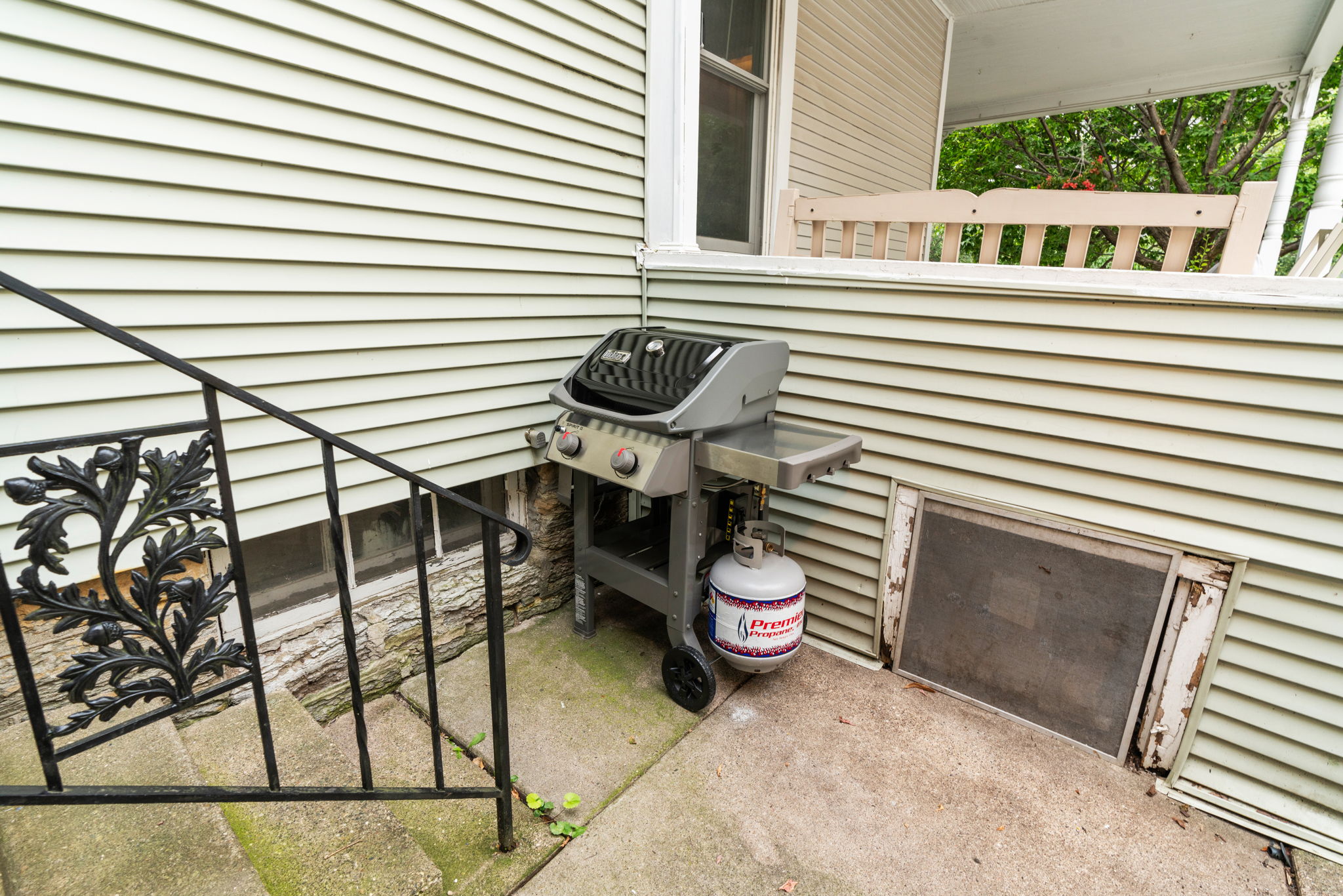 A modern gas grill set against a pale green house with elegant wrought iron railings.