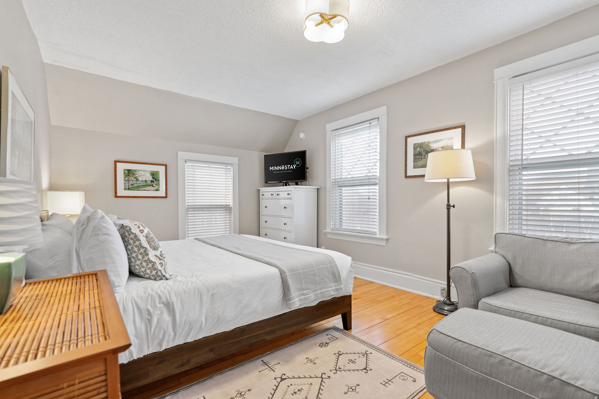 A cozy bedroom featuring a queen bed, gray couch, and bright natural light.