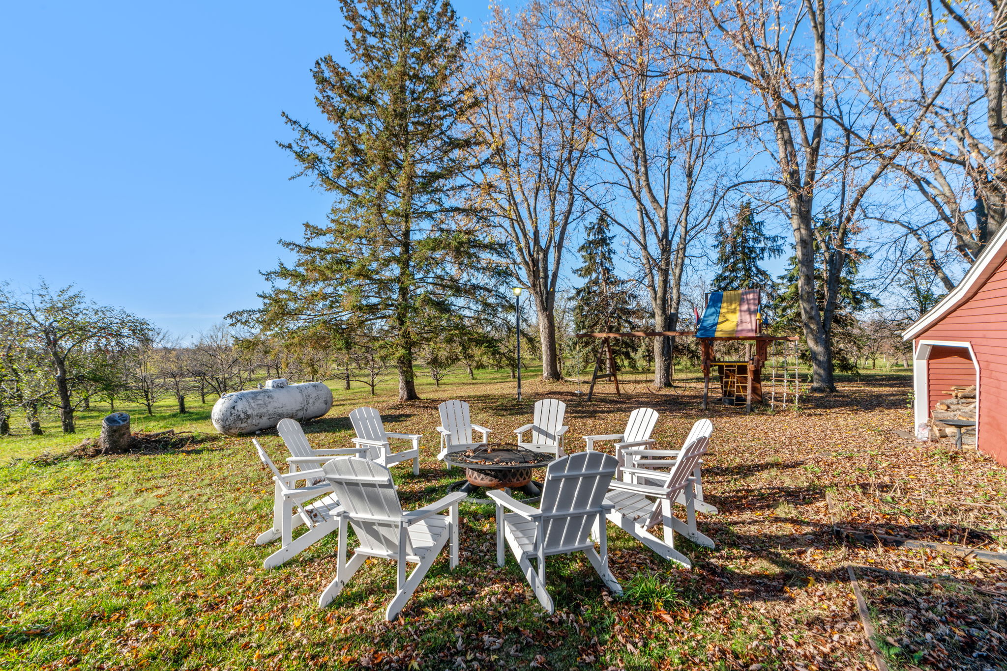 A cozy backyard with Adirondack chairs around a fire pit, surrounded by autumn leaves and a play structure in the trees.