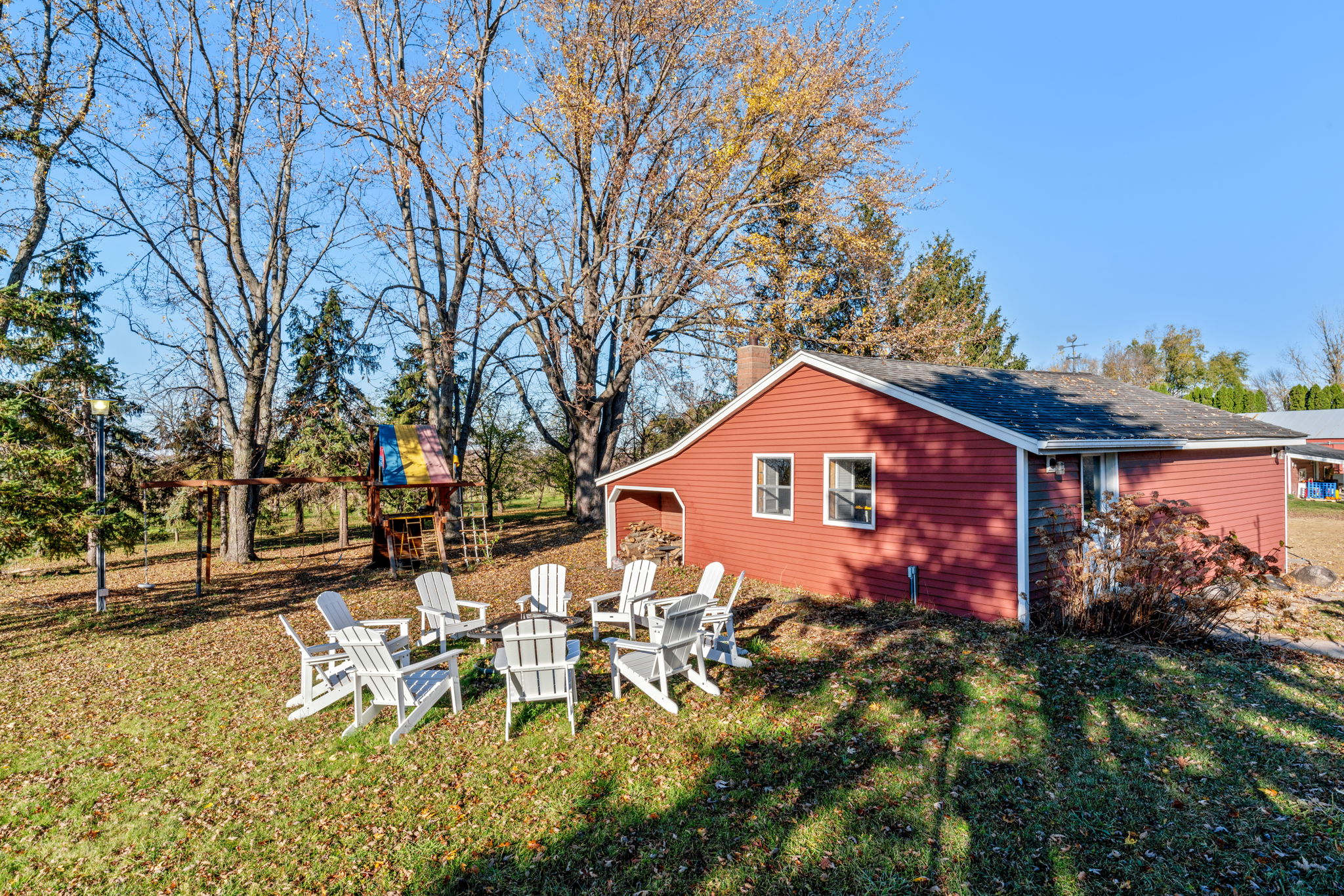A cozy red home with an inviting backyard space featuring white Adirondack chairs and a colorful playhouse.