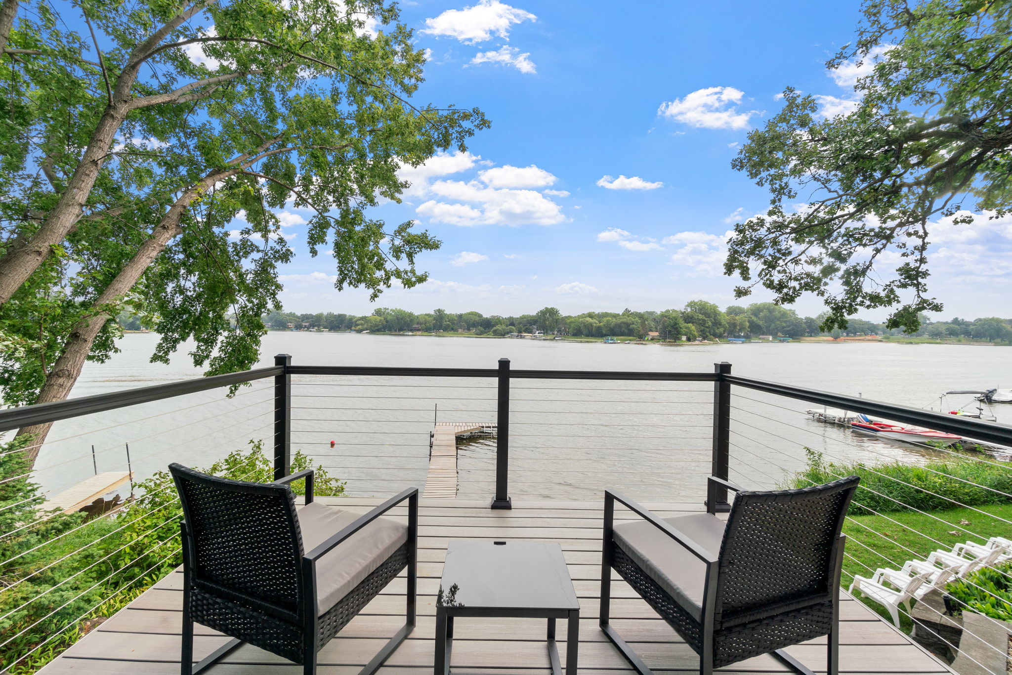 A tranquil lakeside view from a cozy balcony, complete with black wicker chairs and a small table.