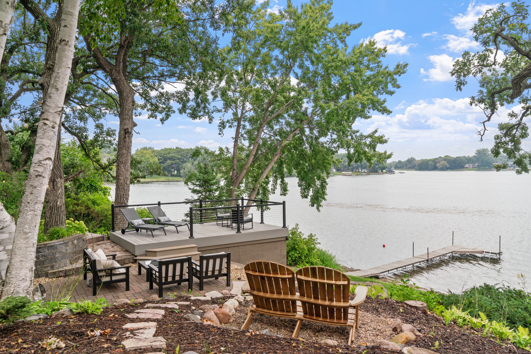 Outdoor seating area by a lake with Adirondack chairs and a dock in the background.