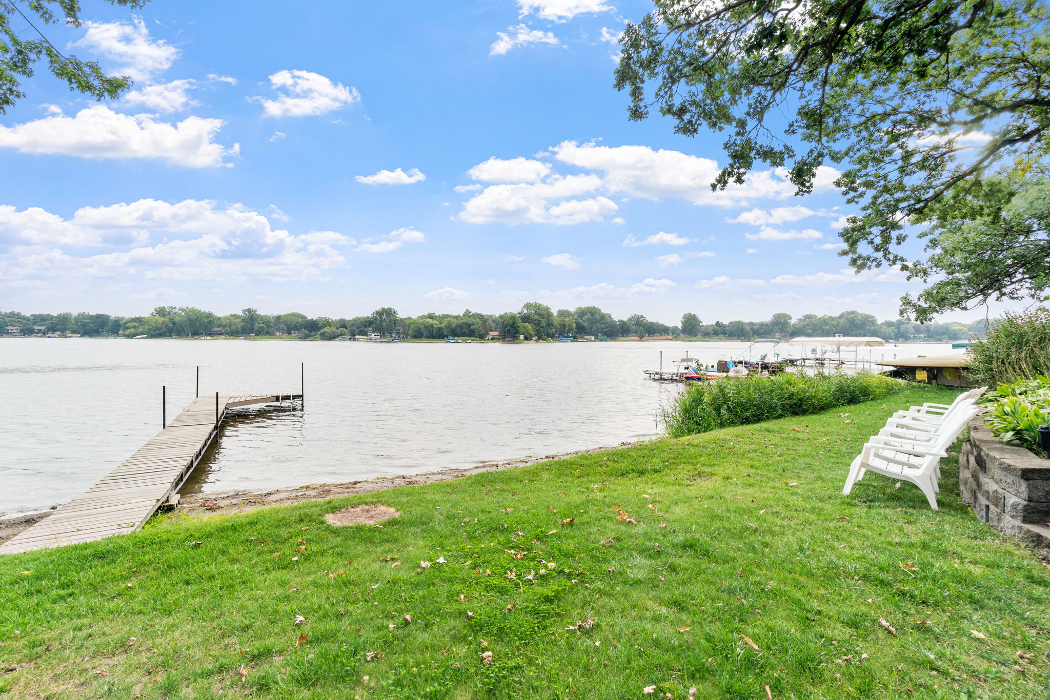 Tranquil lakeside view with a wooden dock and white lawn chairs.