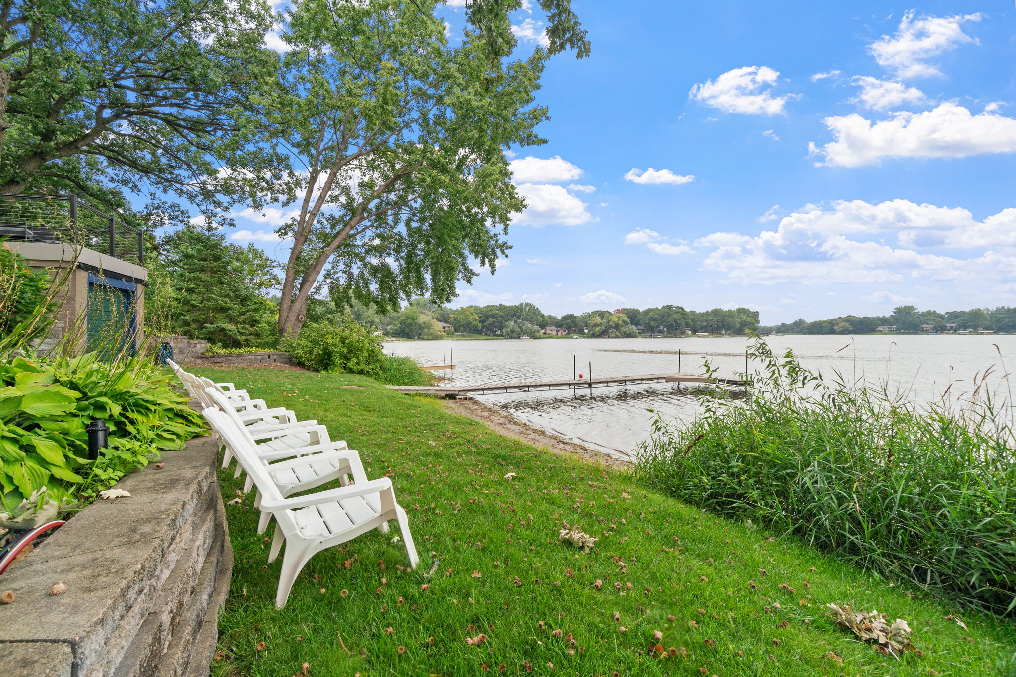 A tranquil lakeside view with white chairs and a wooden dock under a clear blue sky.