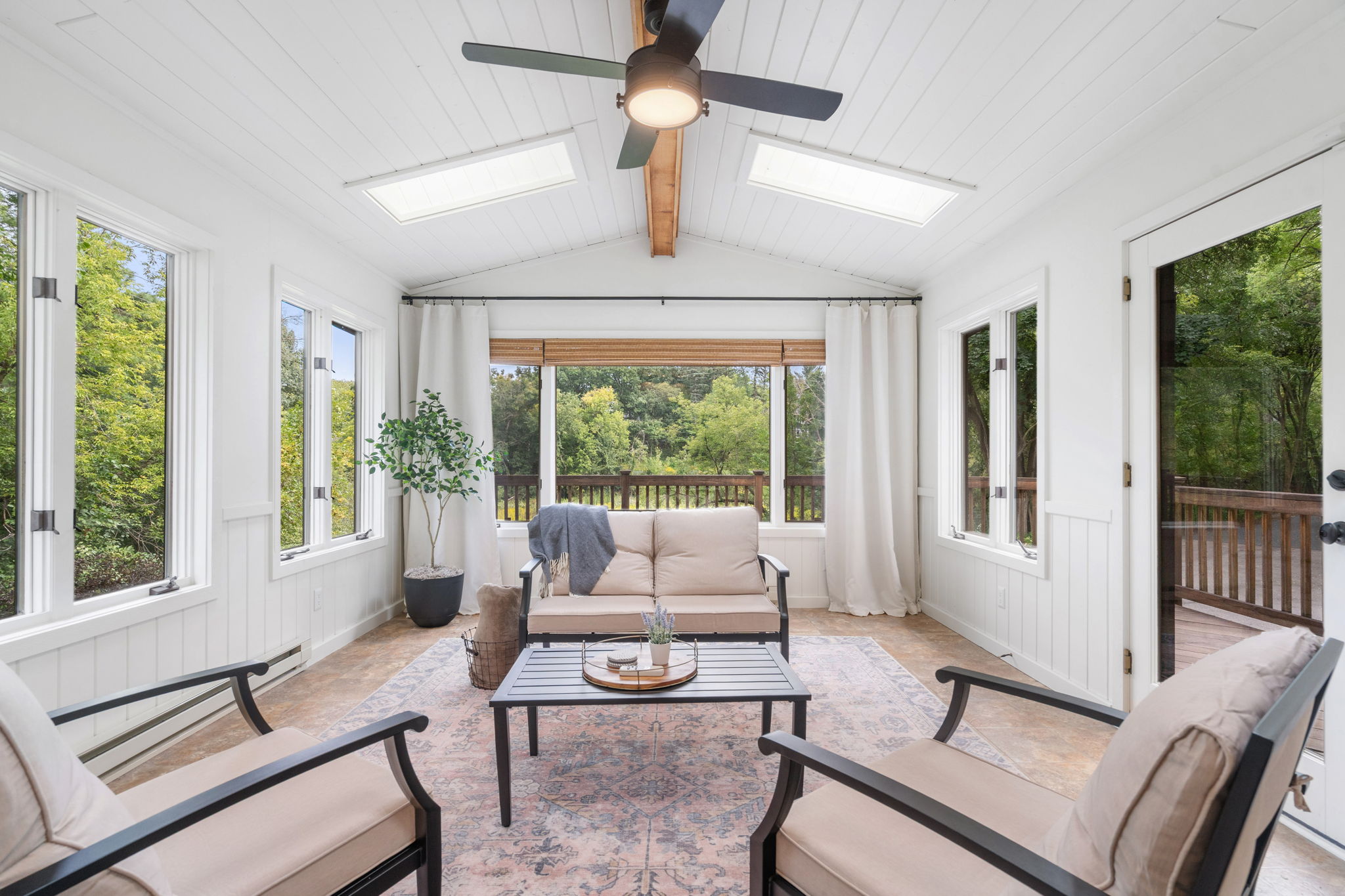 A sunroom with beige sofas, a coffee table, and large windows showcasing greenery outside.