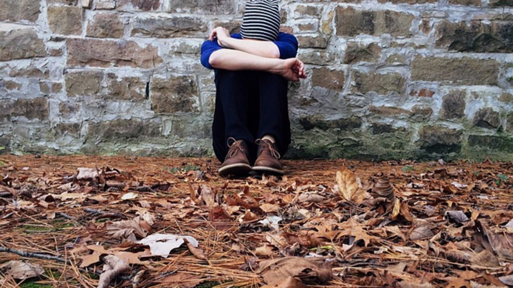 A person sitting quietly against a stone wall among fallen autumn leaves