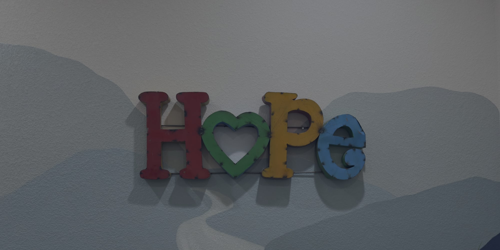 Colorful letters spelling HOPE, with a green heart in place of the O, mounted on a wall painted with soft blue mountain shapes