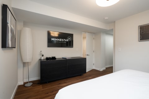 Interior view of a modern vacation rental room with minimalist decor, including a bed, lamp, entertainment unit, and abstract art.