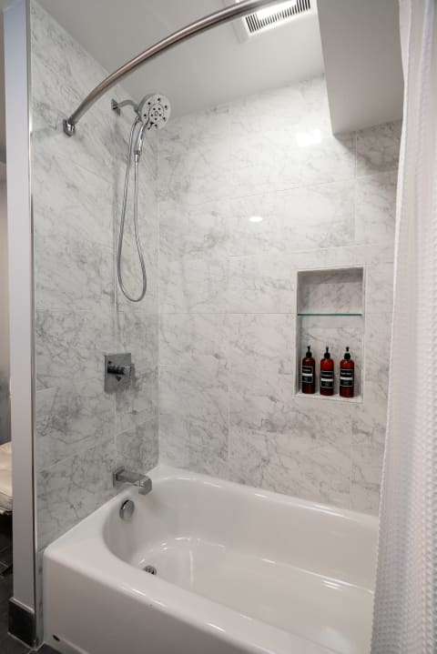 A contemporary shower with marble tiles and a white tub, showcasing bath products on a shelf.