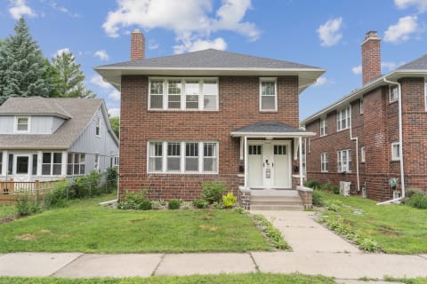 A two-story red brick building with double doors and windows, next to a smaller blue house.
