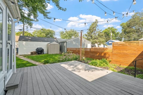 View of a backyard featuring a wooden deck, grass, storage sheds, and landscape elements.