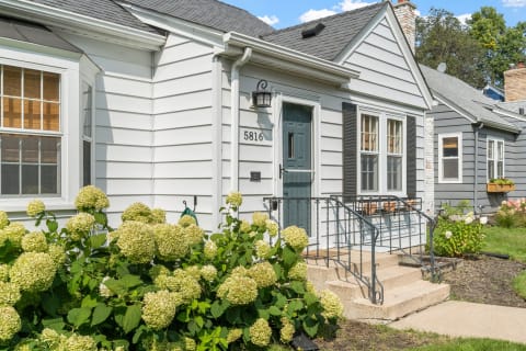 Exterior view of a house with a blue front door, iron railing, and flowering bushes.