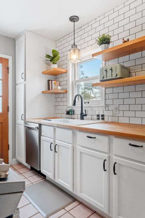 A bright kitchen with white cabinets, wooden countertops, and open shelves containing plants and cookbooks.