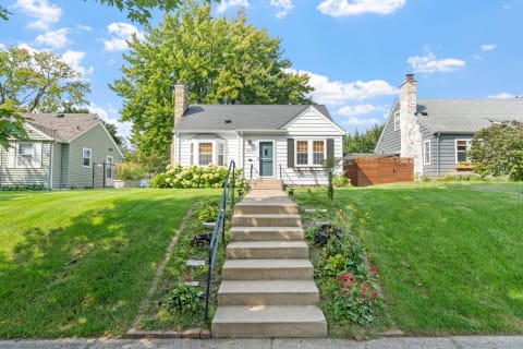 A single-story house with white siding, blue door, and lush green lawn surrounded by flowers.