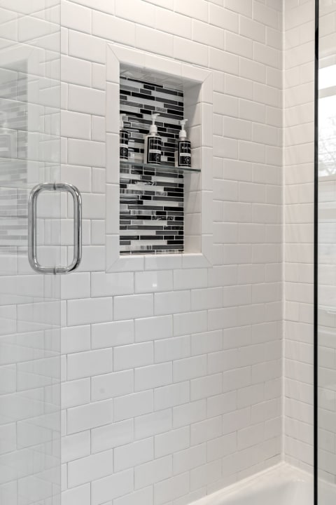 A modern shower with white subway tiles, a glass panel, and a niche holding black bottles against a black and silver mosaic.